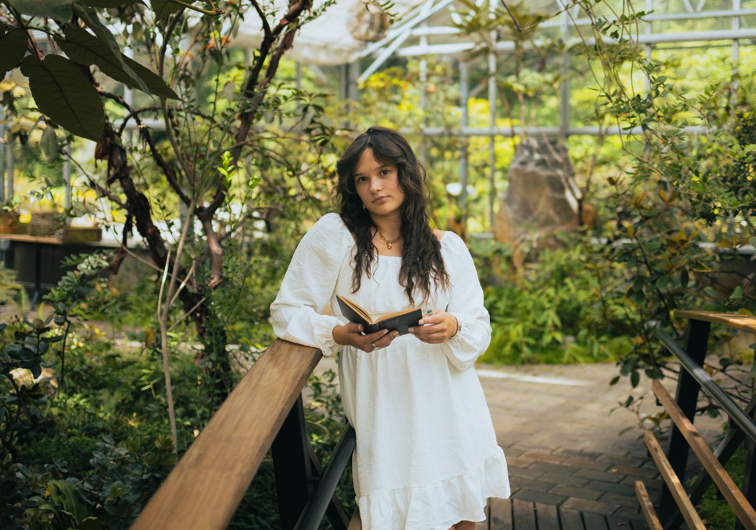 Young woman in a white dress holds an open book while standing on a wooden walkway in a lush garden greenhouse.