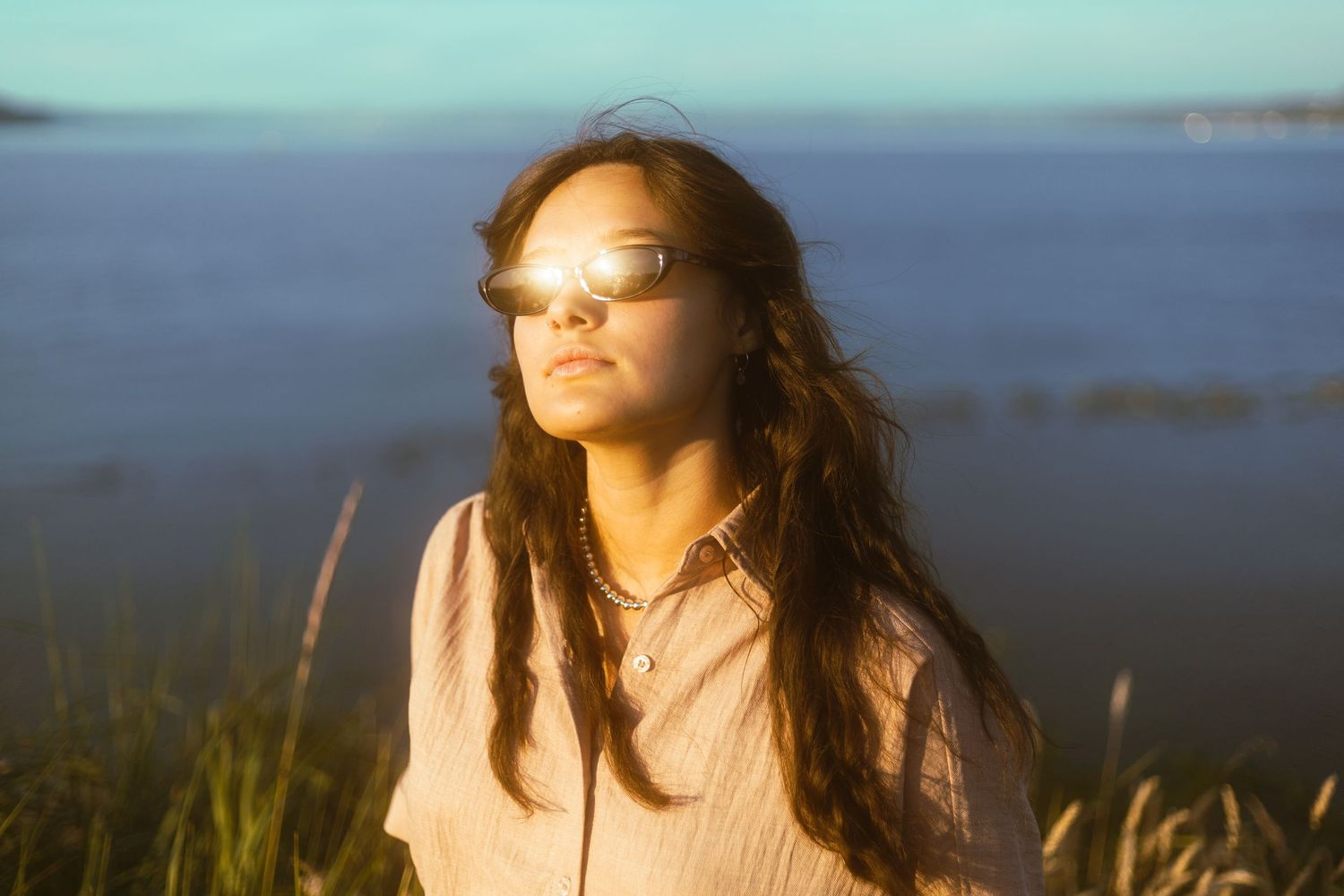 Young woman with sunglasses enjoys golden hour sunlight near a tranquil blue lake outdoors.