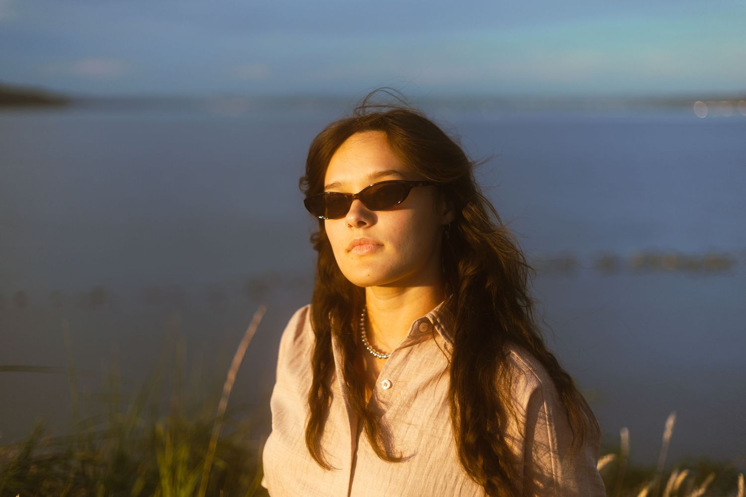 Young woman with long dark hair wearing sunglasses poses outdoors near a calm waterway in warm golden sunset light.