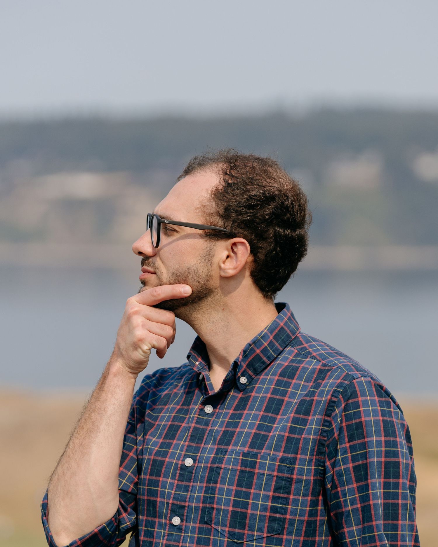 Thoughtful man with glasses in plaid shirt gazes into distance near waterfront.