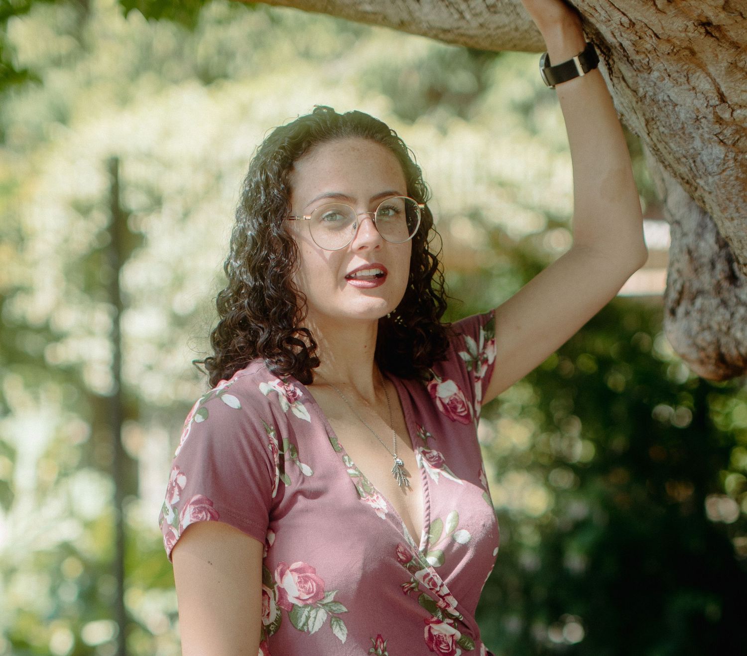 Young woman in floral dress and glasses leans against tree in sunny outdoor garden setting.
