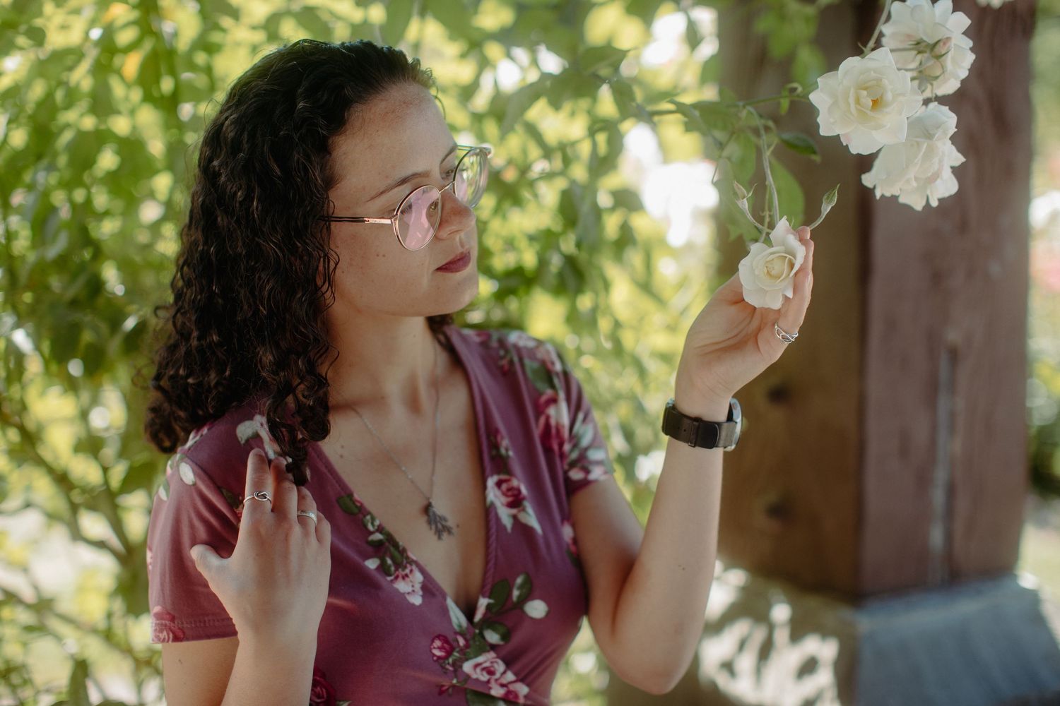 A young woman with curly hair and glasses in a pink floral dress examines white roses in a garden.