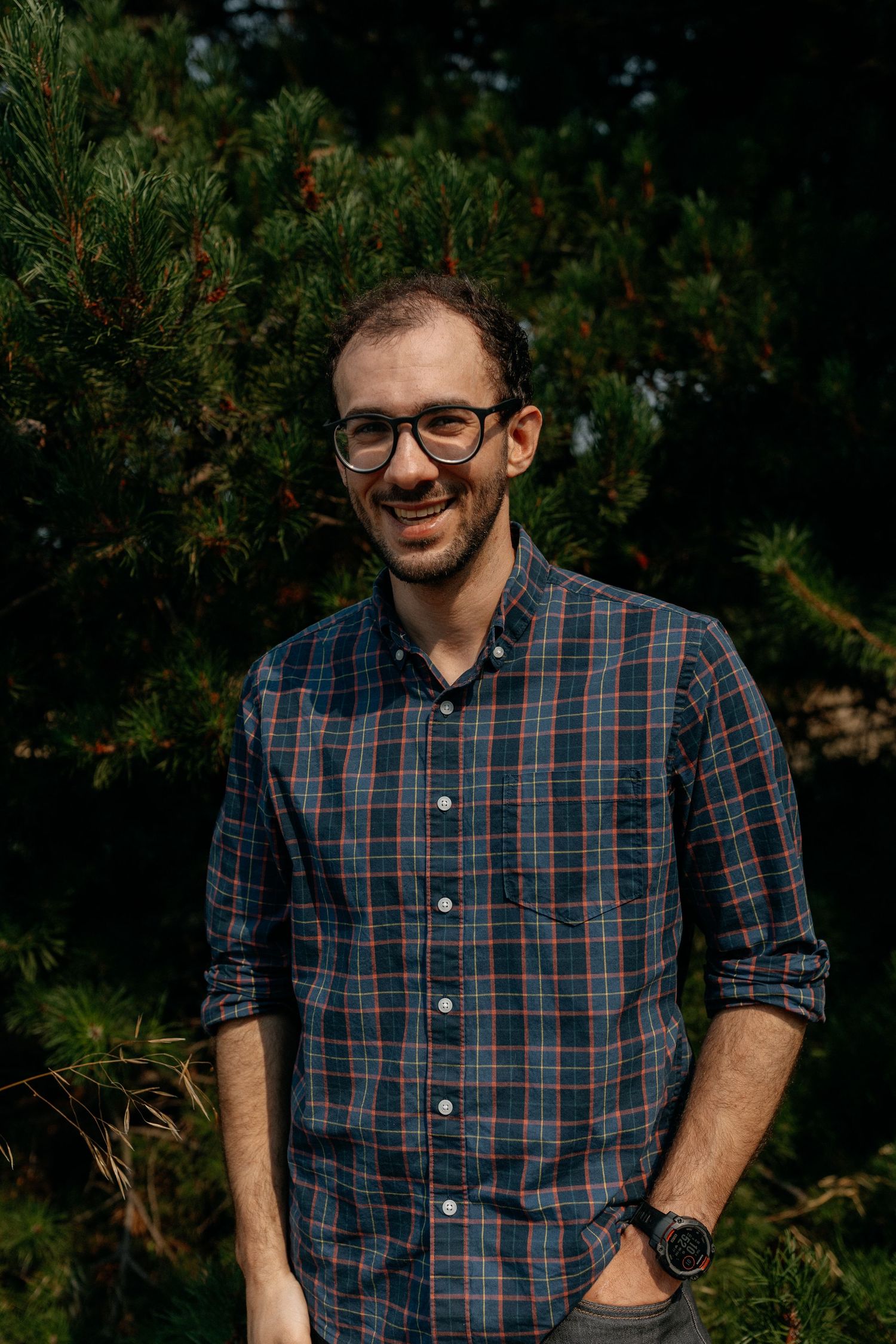 Cheerful man with glasses in blue plaid shirt smiling outdoors near evergreen trees.
