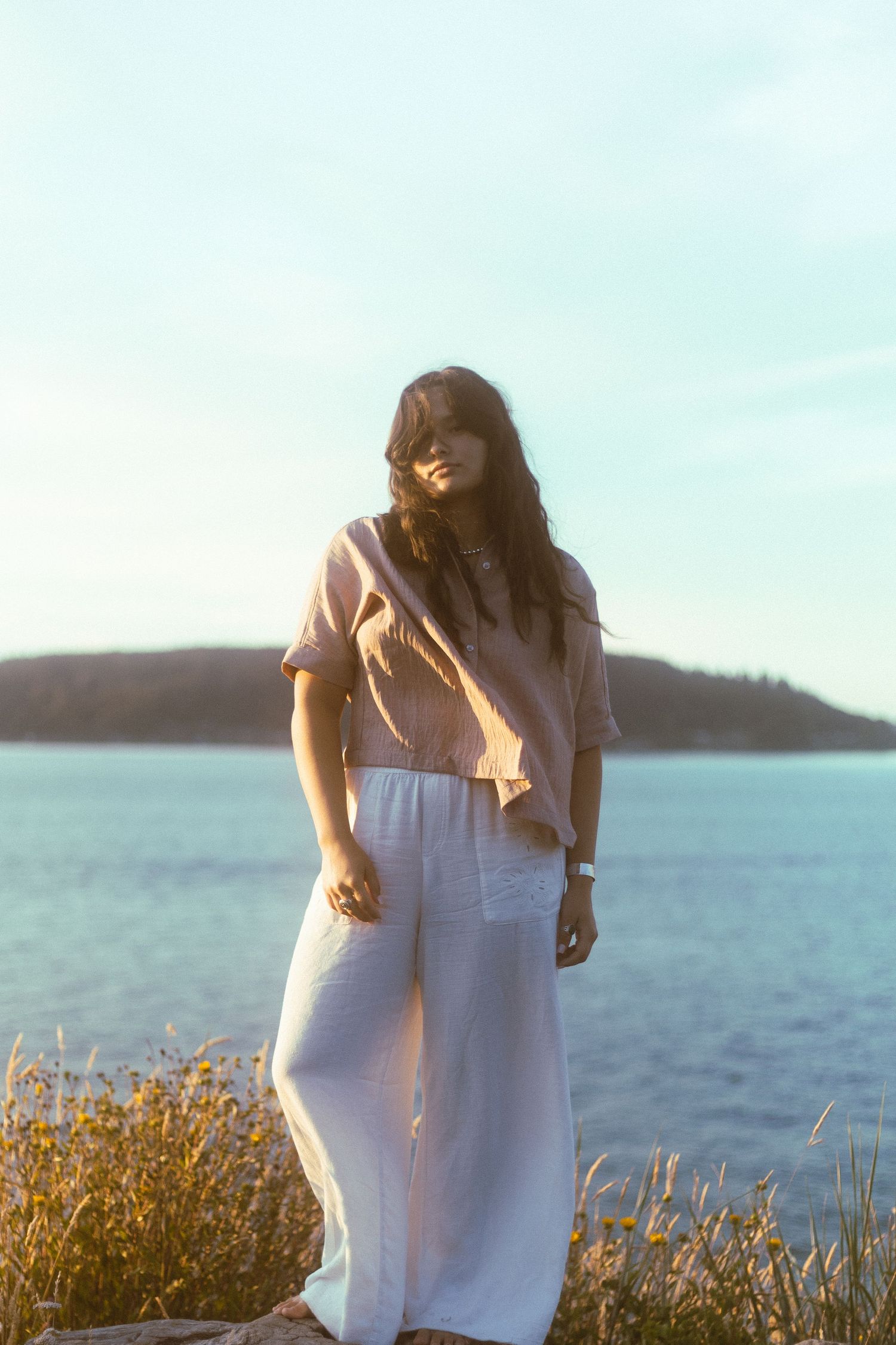 Young woman in white pants and pink top stands on rocky shore at golden hour sunset.