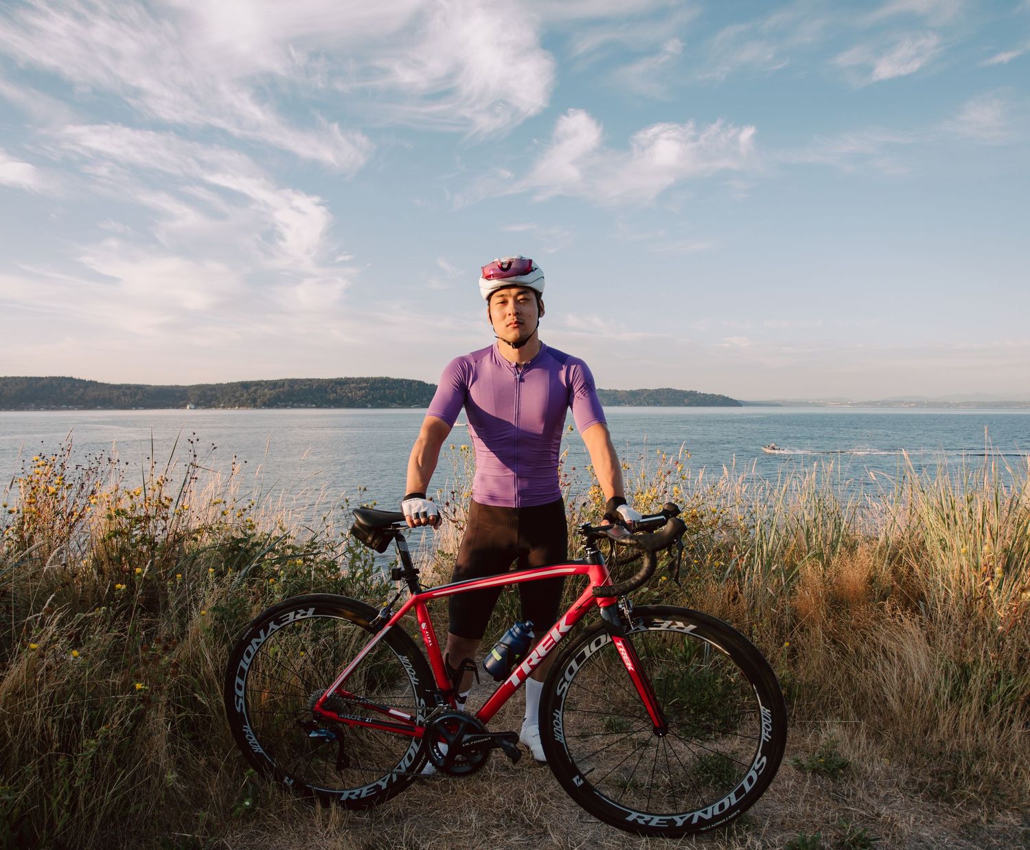 Cyclist in purple jersey standing with red road bike on coastal trail with scenic ocean and cloudy sky behind him.
