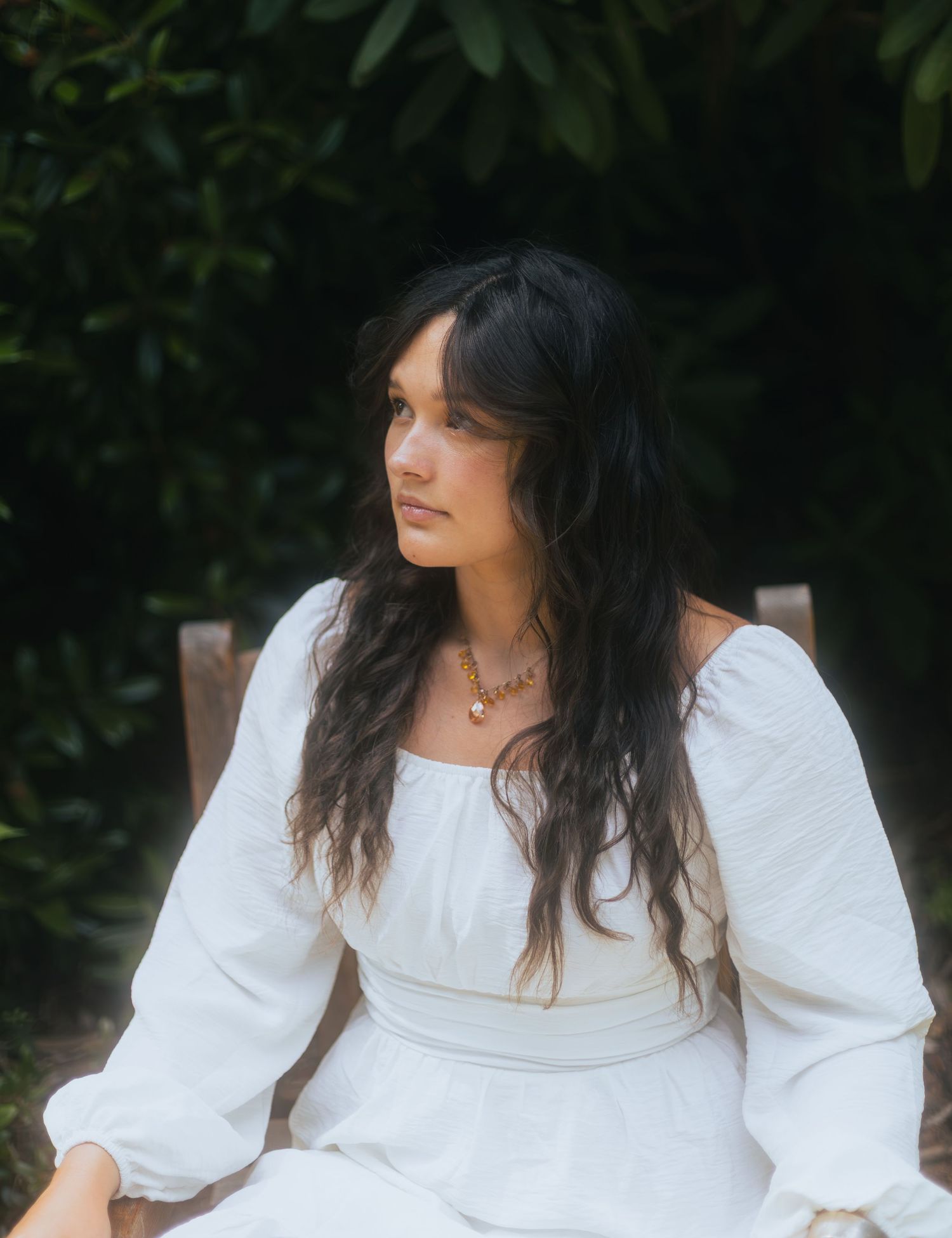 Woman with long dark wavy hair in white off-shoulder dress sitting outdoors near green shrubs.