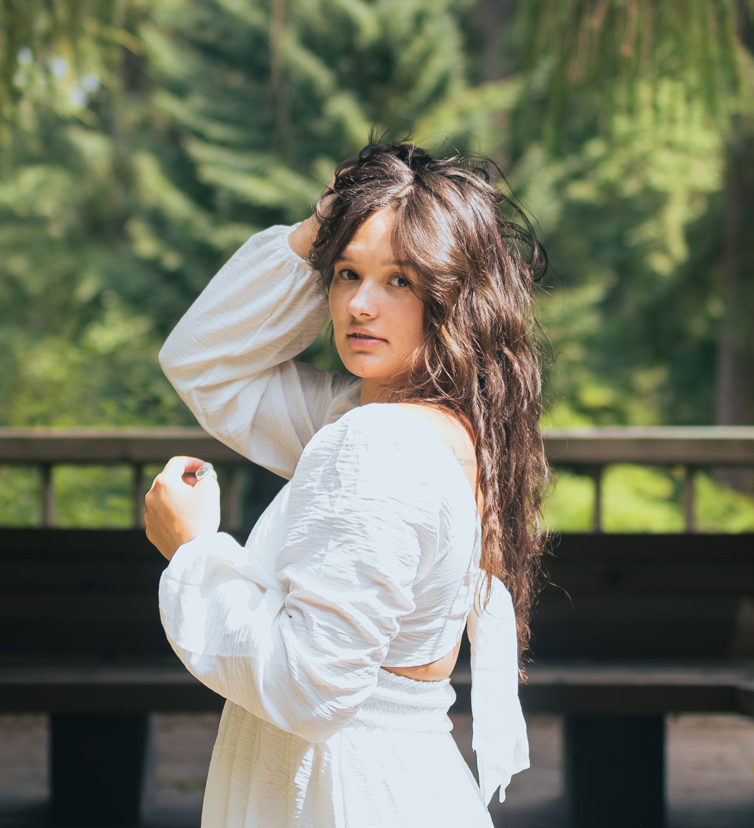 Woman in white backless dress posing outdoors near wooden railing with pine trees behind her.