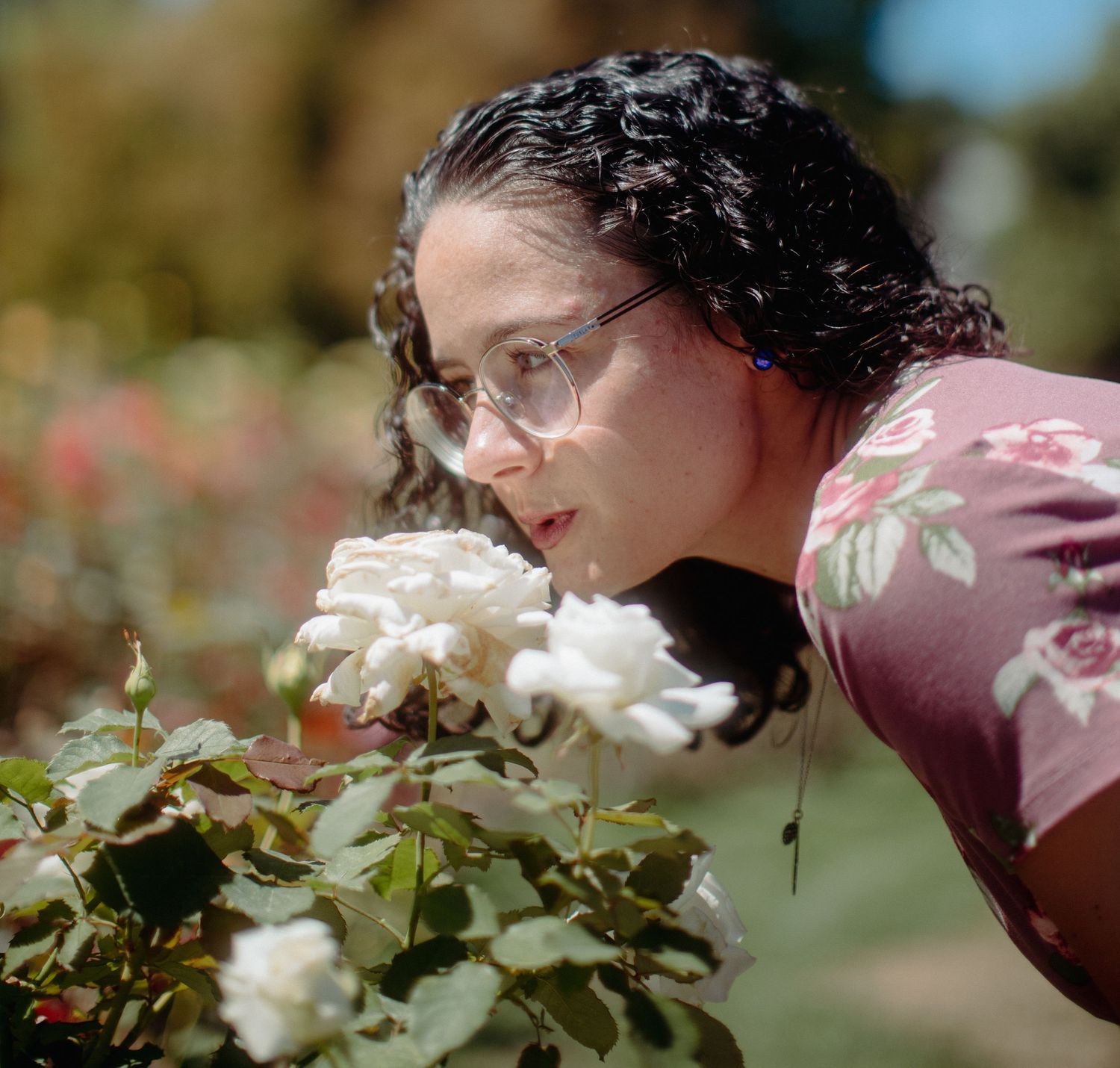 A young woman with curly dark hair and glasses leans in to smell white roses in a sunny garden.