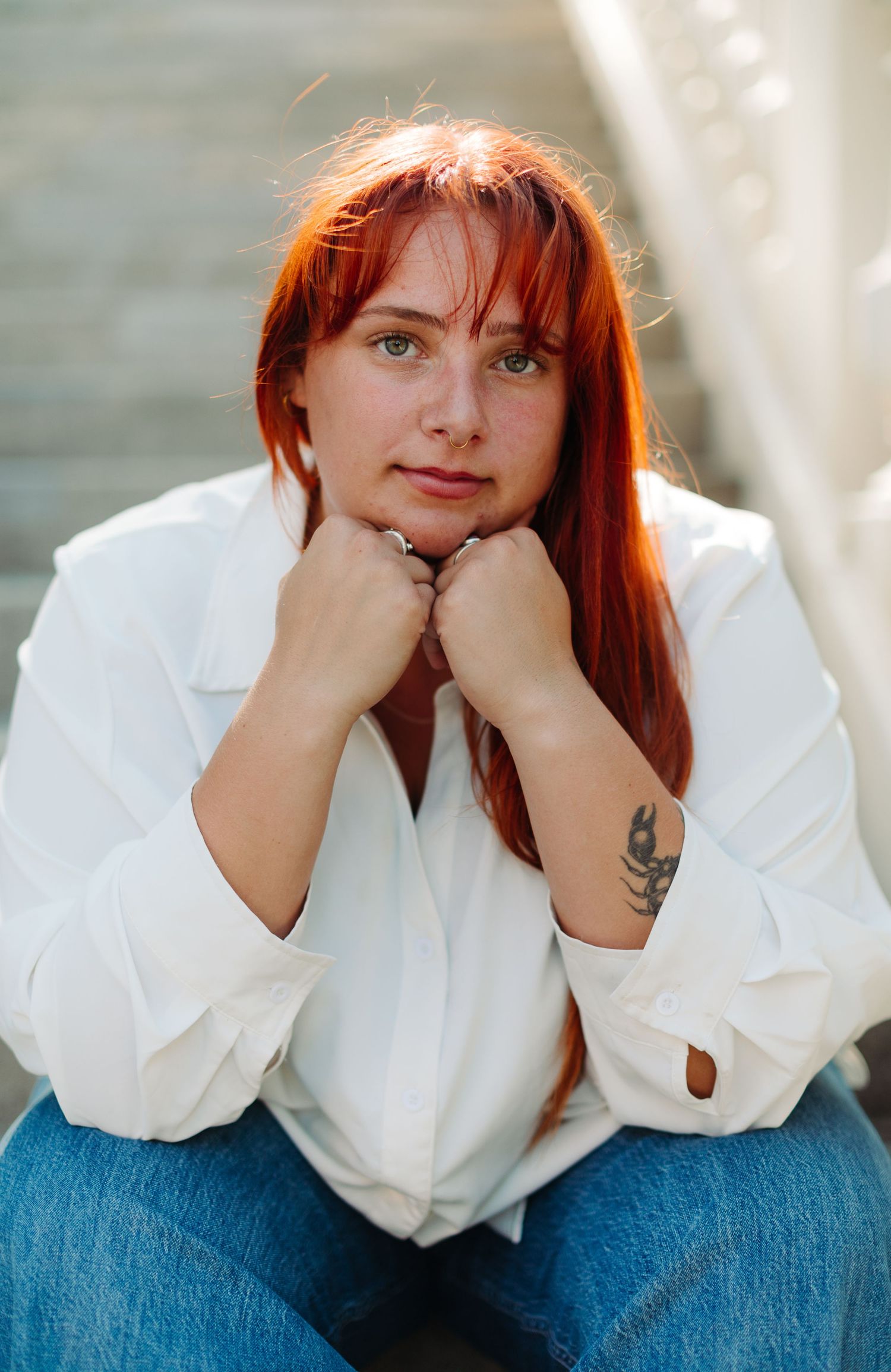 Red-haired woman with green eyes and arm tattoo sitting on steps wearing white shirt and jeans.