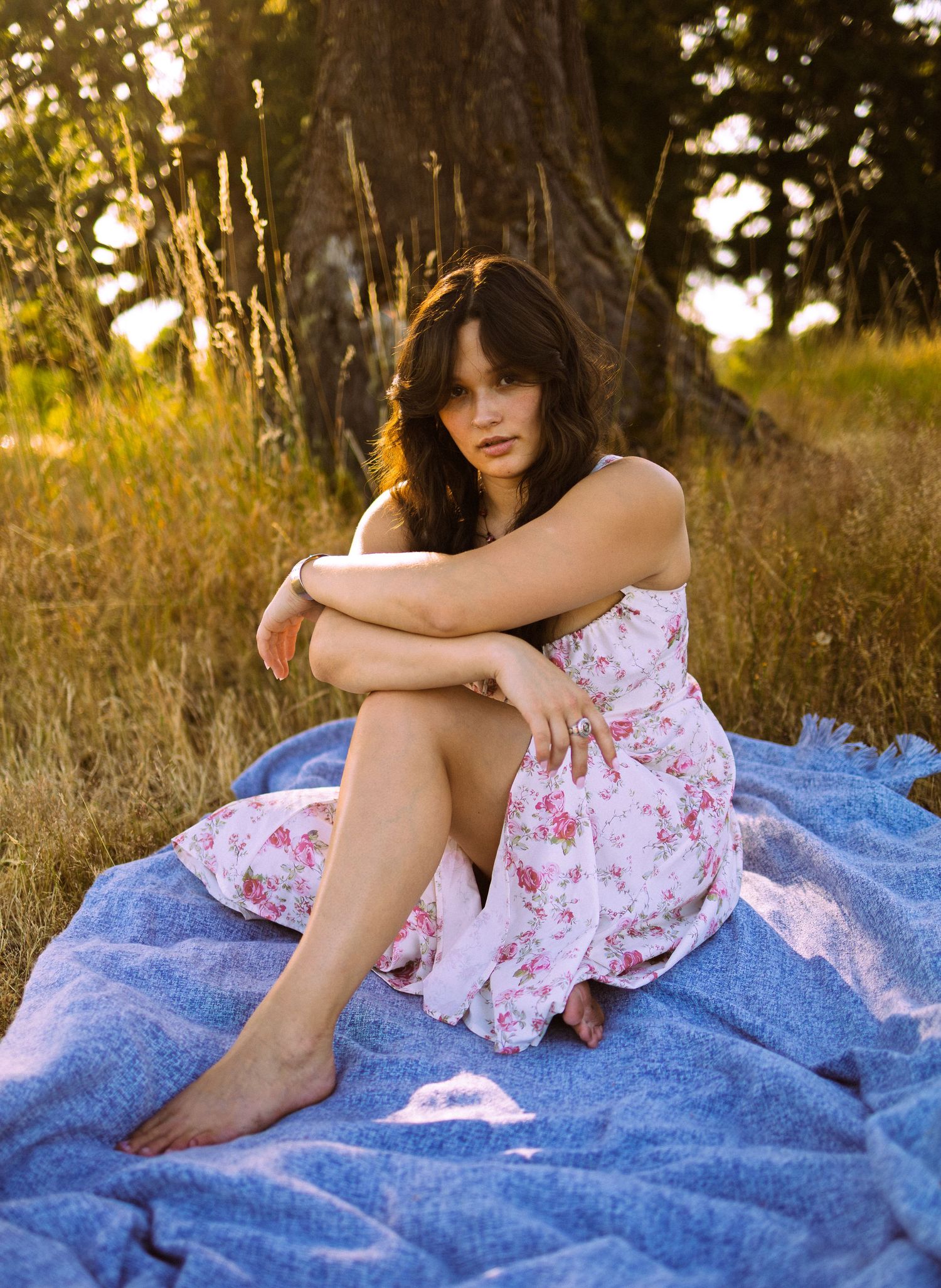 Young brunette woman in floral dress sits barefoot on blanket in sunlit meadow near trees.