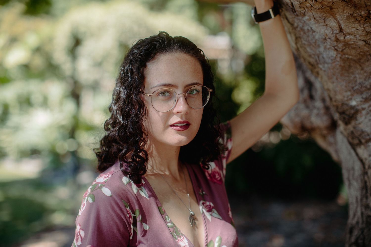 Curly-haired woman in floral dress and glasses leans against tree in sunny park setting.
