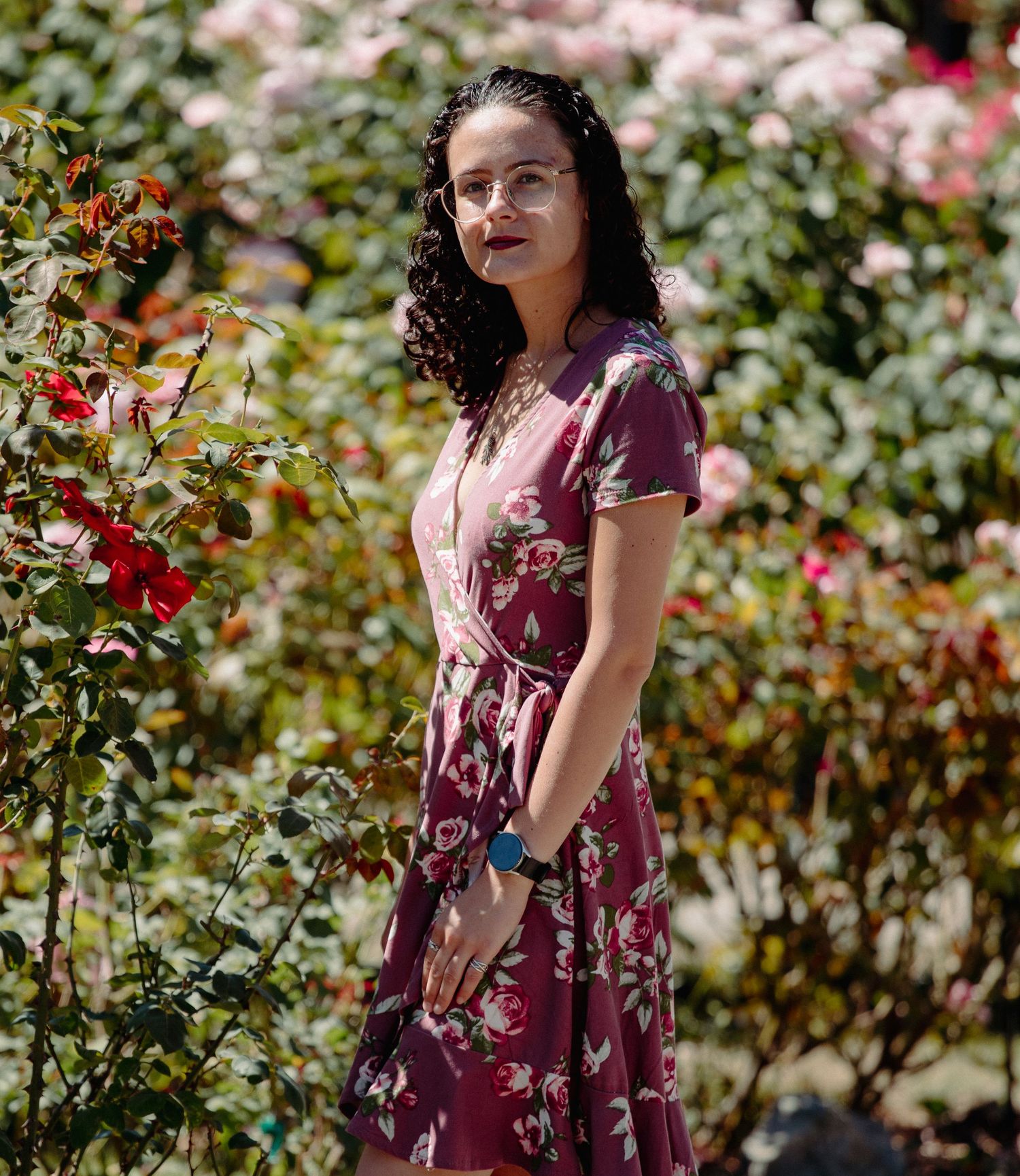 A young woman with curly hair wearing glasses and a purple floral dress stands among colorful roses in a garden.