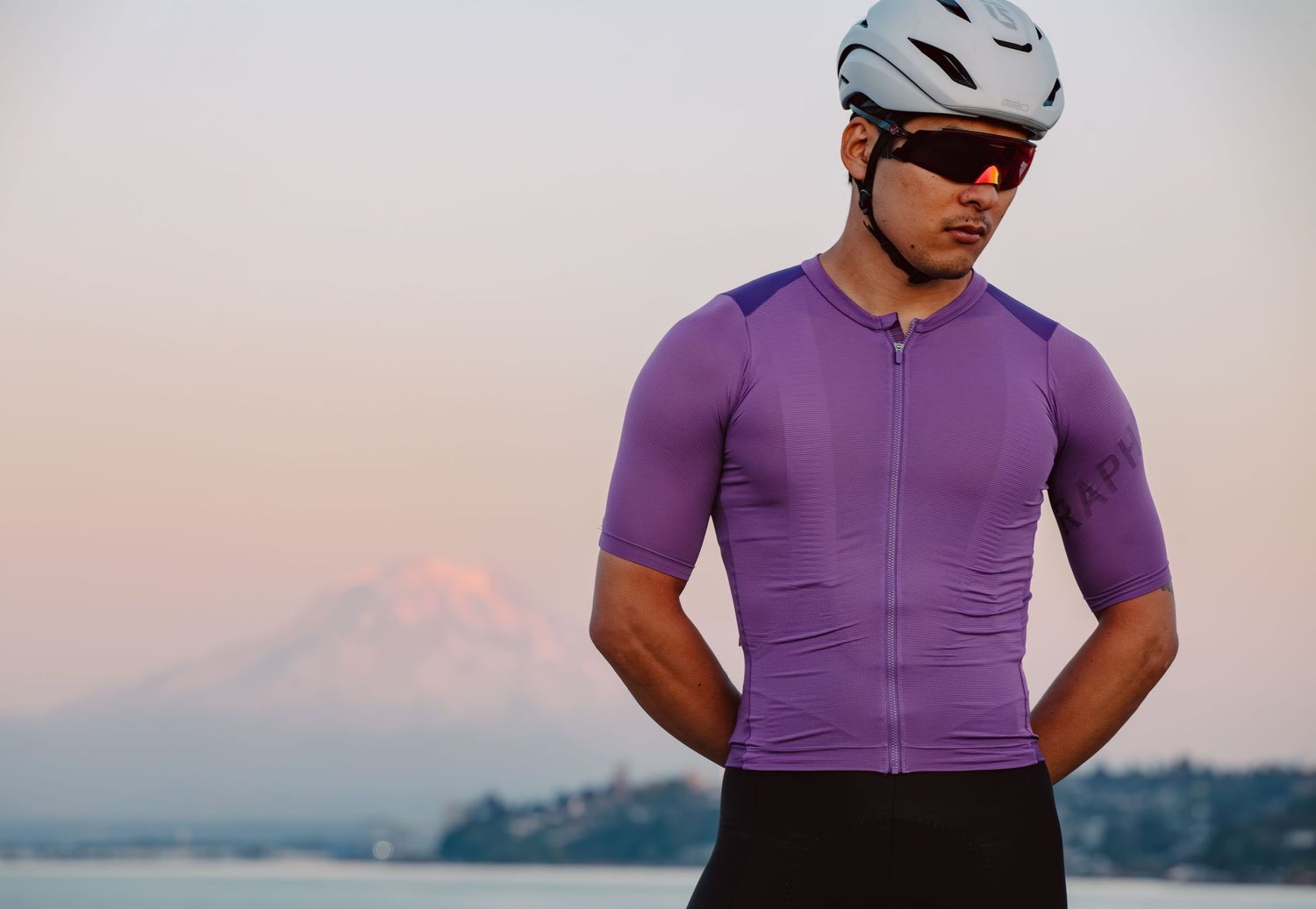 Male cyclist in purple jersey and white helmet stands with mountain and water backdrop.