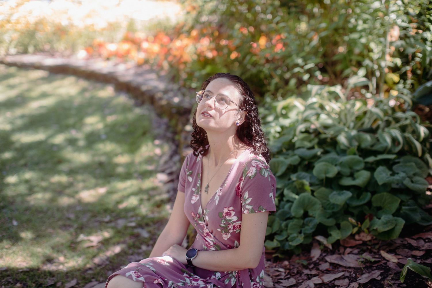 A young woman with curly hair in a pink floral dress sits in a sunny garden with eyes closed peacefully.