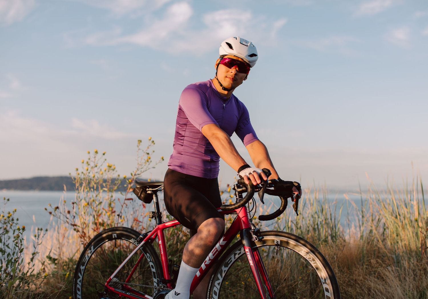 Male cyclist in purple jersey and white helmet resting on red road bike with ocean and wildflowers in background.