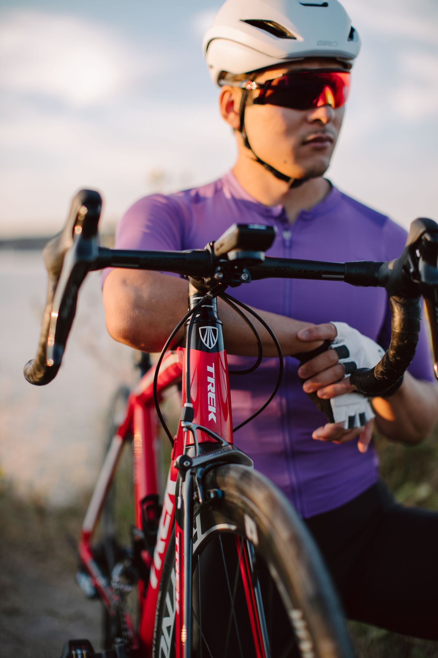 Male cyclist in purple jersey and white helmet with red sunglasses adjusts Trek road bike near a scenic waterfront.