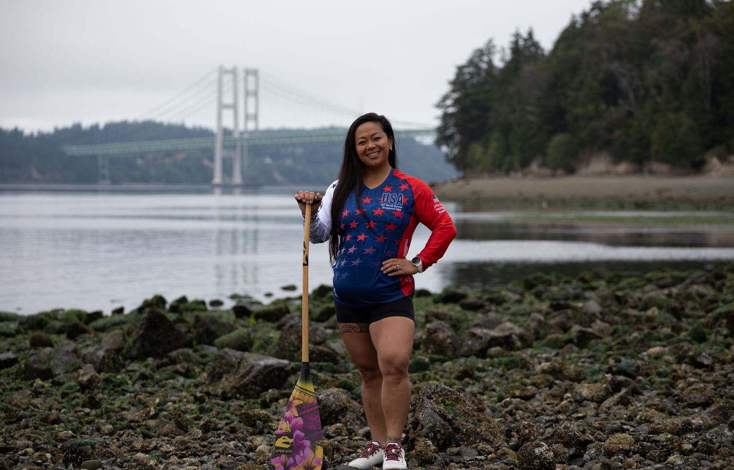 Female athlete in USA uniform poses with a paddle on rocky shoreline with a large suspension bridge in background.