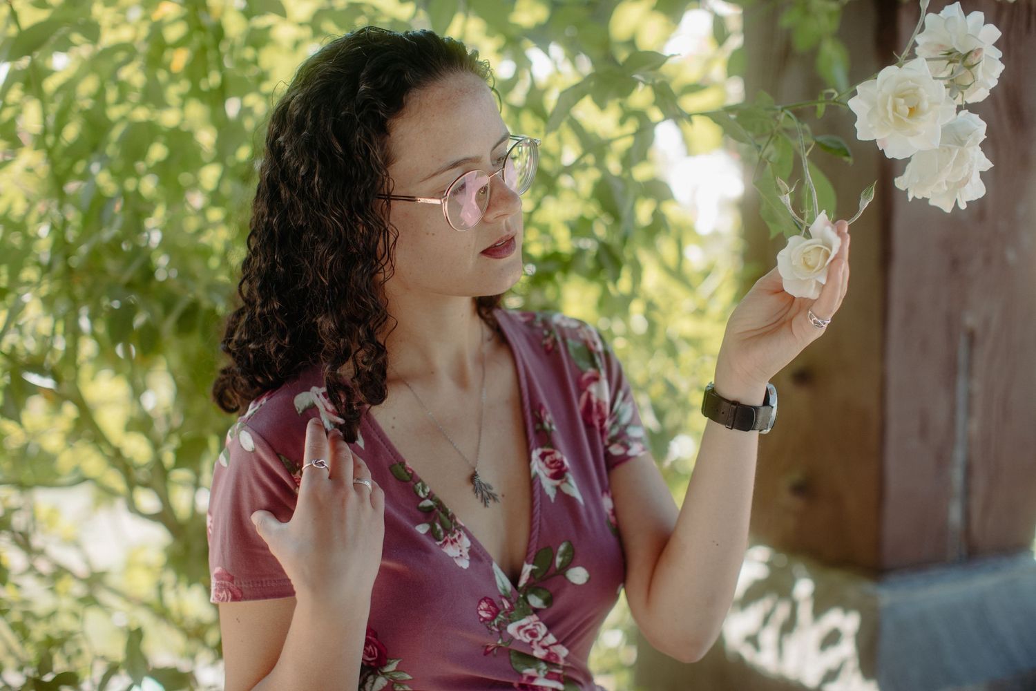 Young woman in floral dress and glasses admires a white flower in a lush garden.