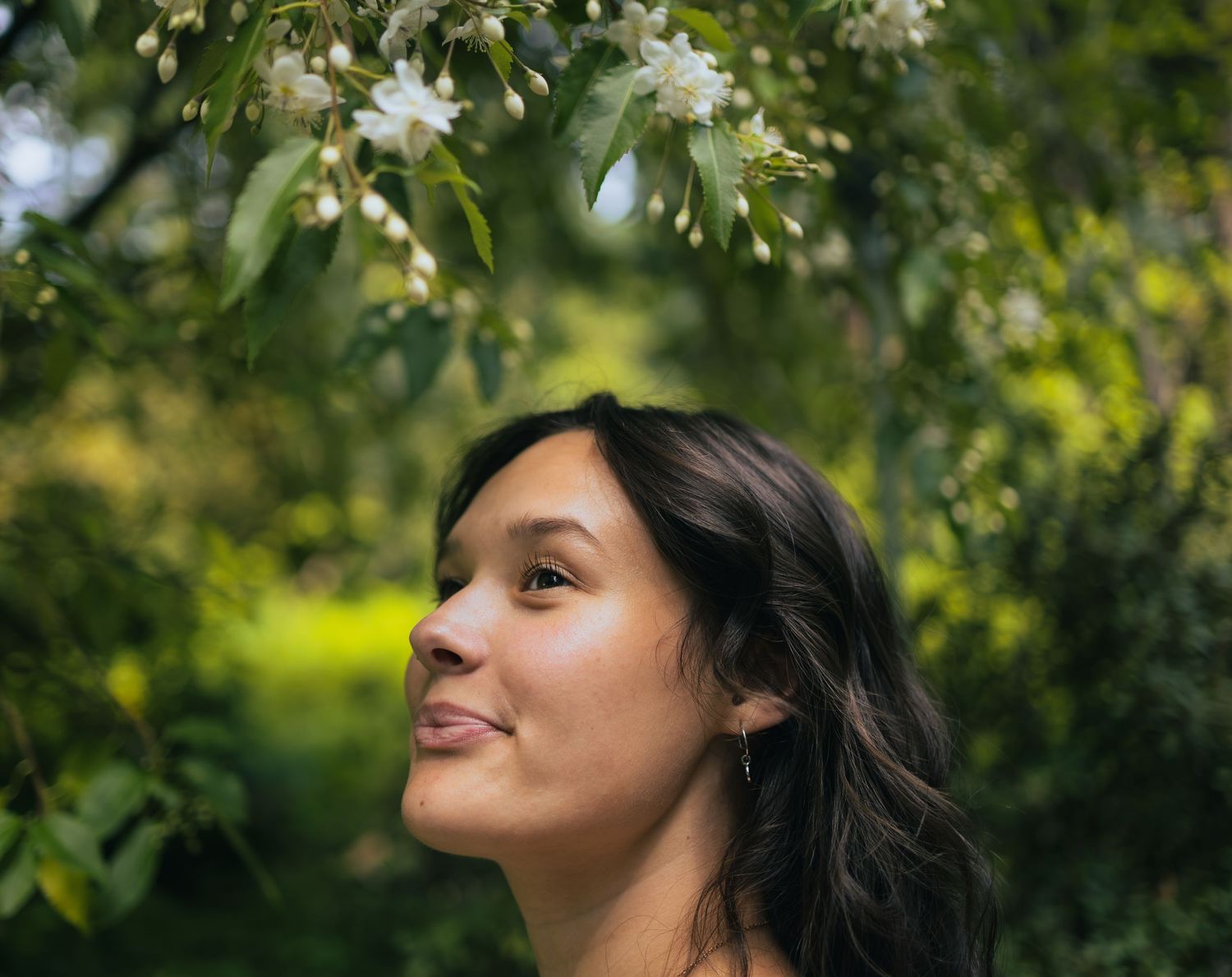Young woman with dark hair smiling upward at white flowering tree branches in garden.