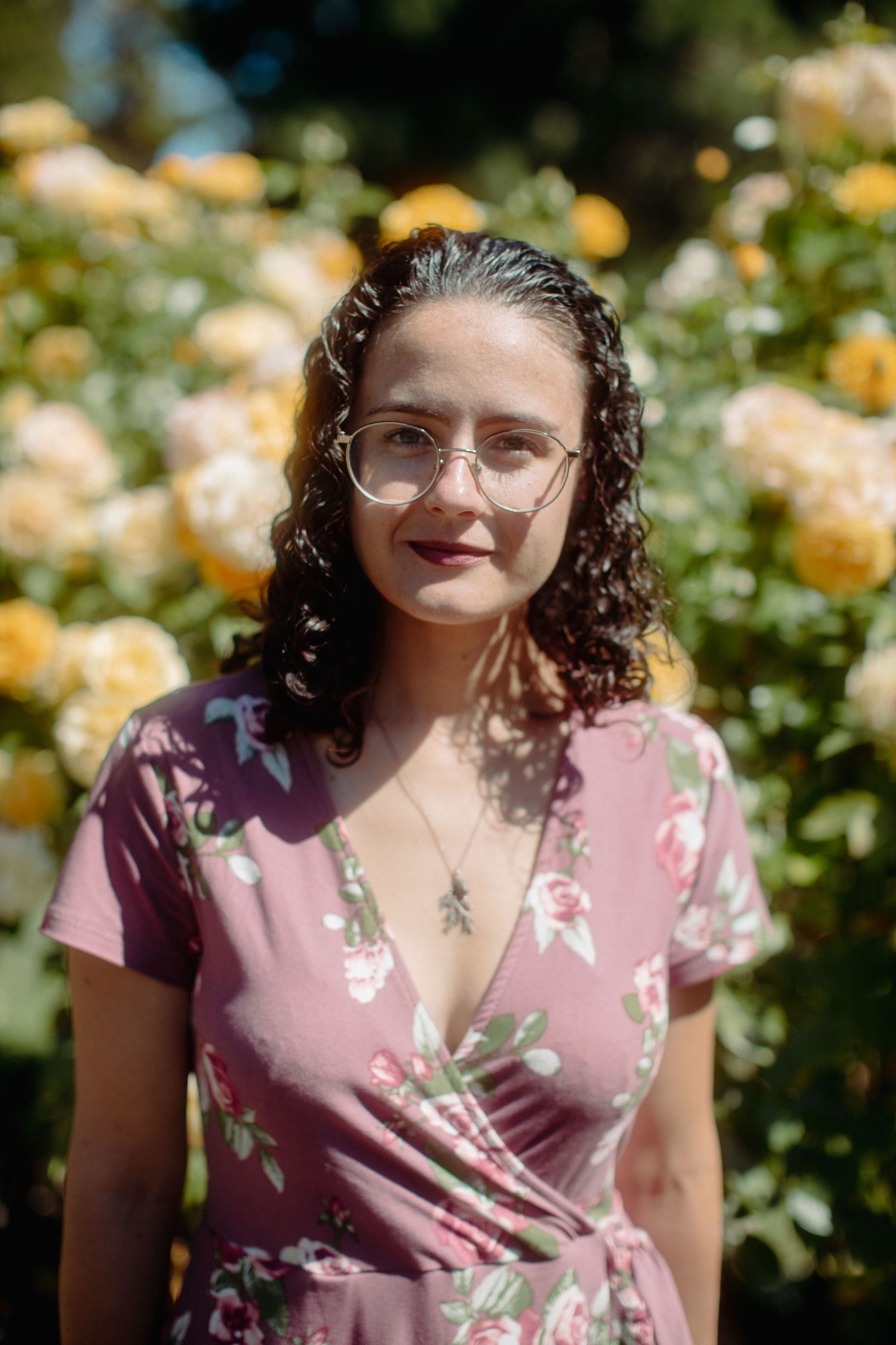 Young woman in pink floral dress and glasses stands smiling before yellow roses in sunlight.