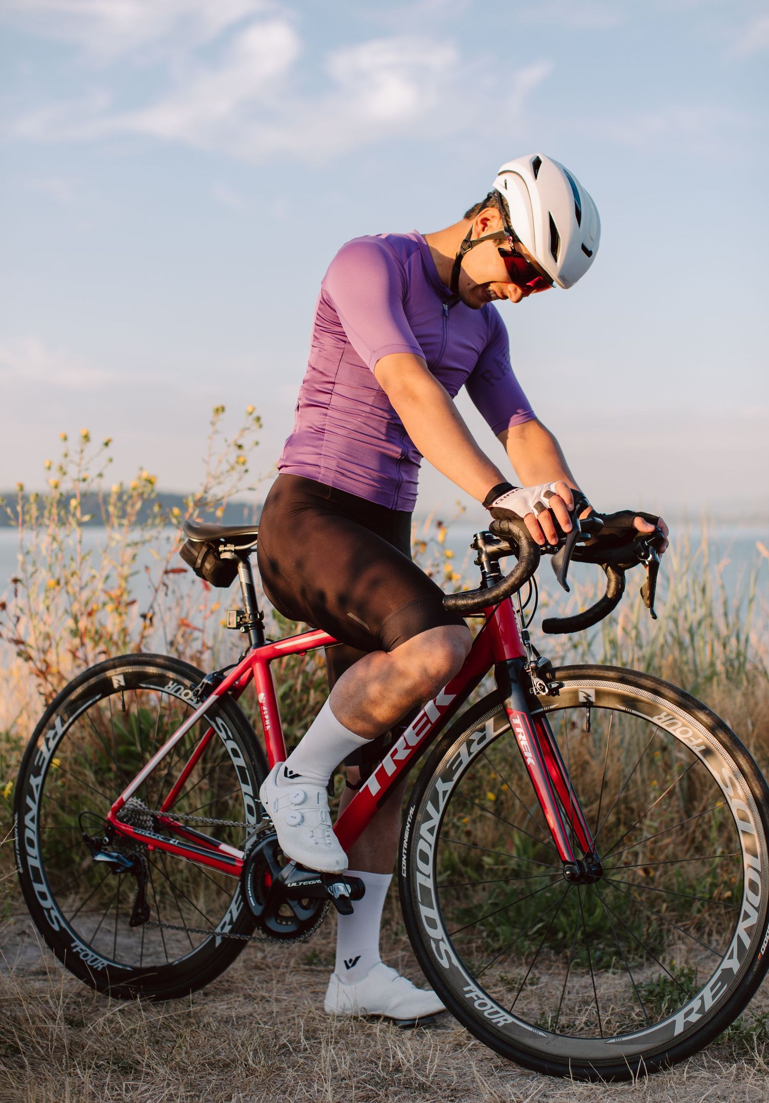 Male cyclist in purple jersey rides red Trek road bike along scenic waterfront trail.
