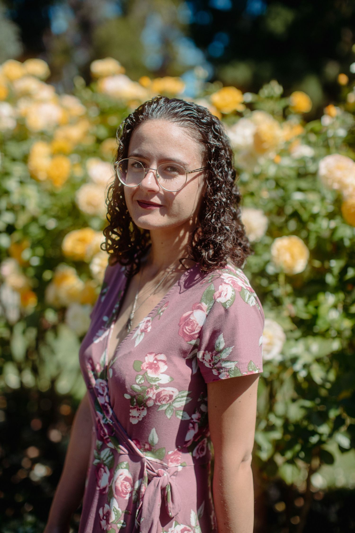 Young woman in floral wrap dress and glasses poses before blooming yellow roses outdoors.