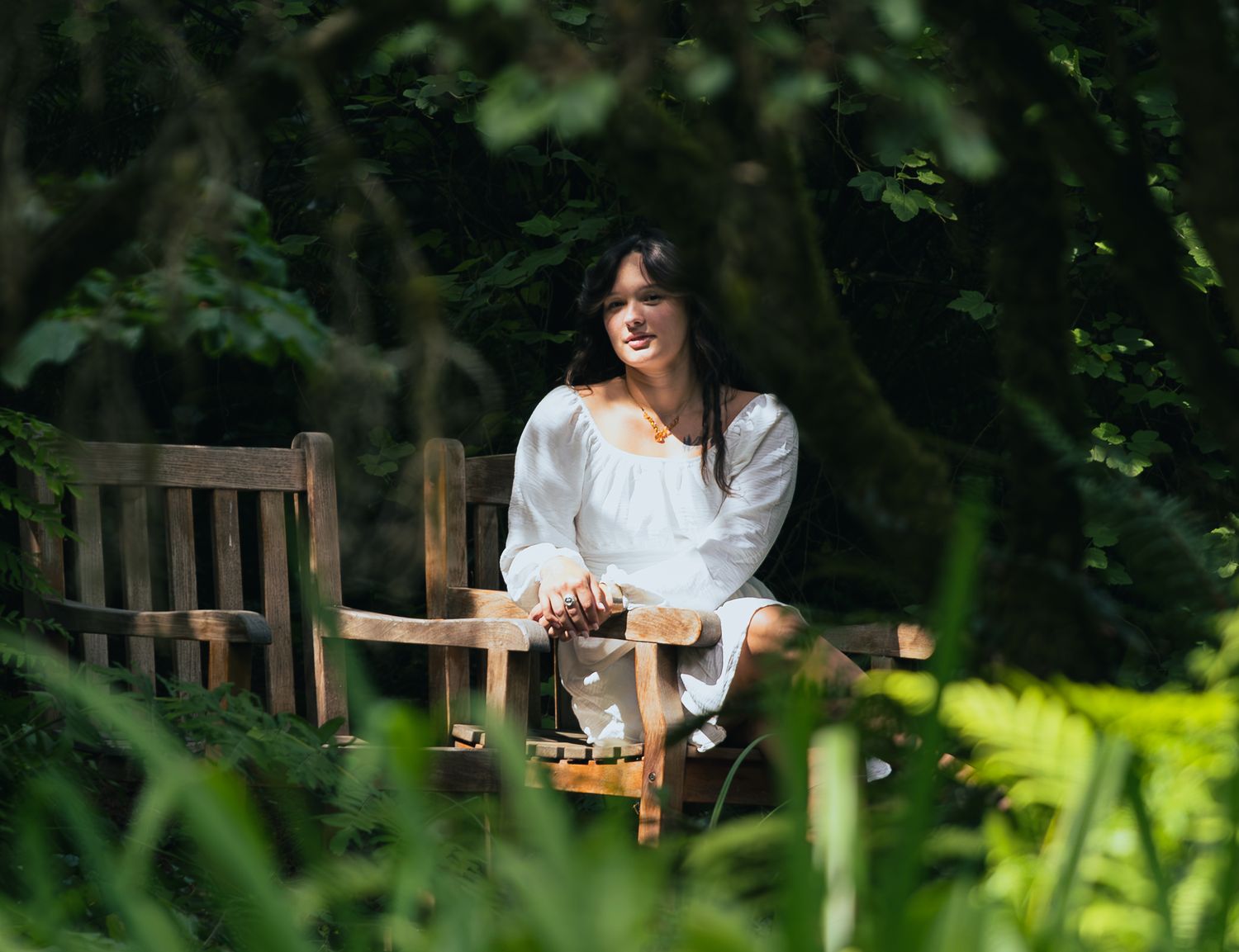 Woman in white dress sitting on wooden bench surrounded by lush green garden foliage.