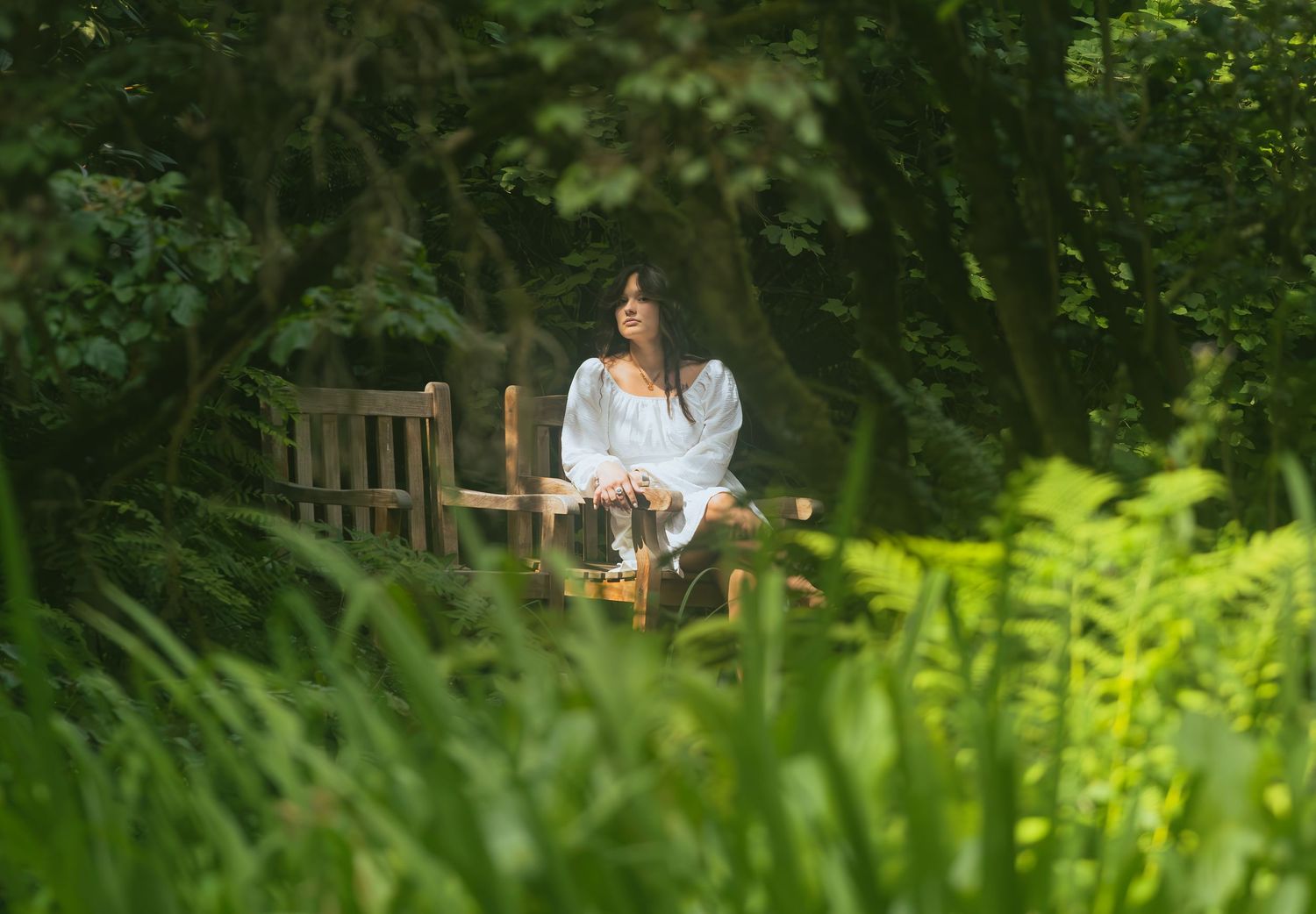 Woman in white dress sits peacefully on bench surrounded by lush green foliage.