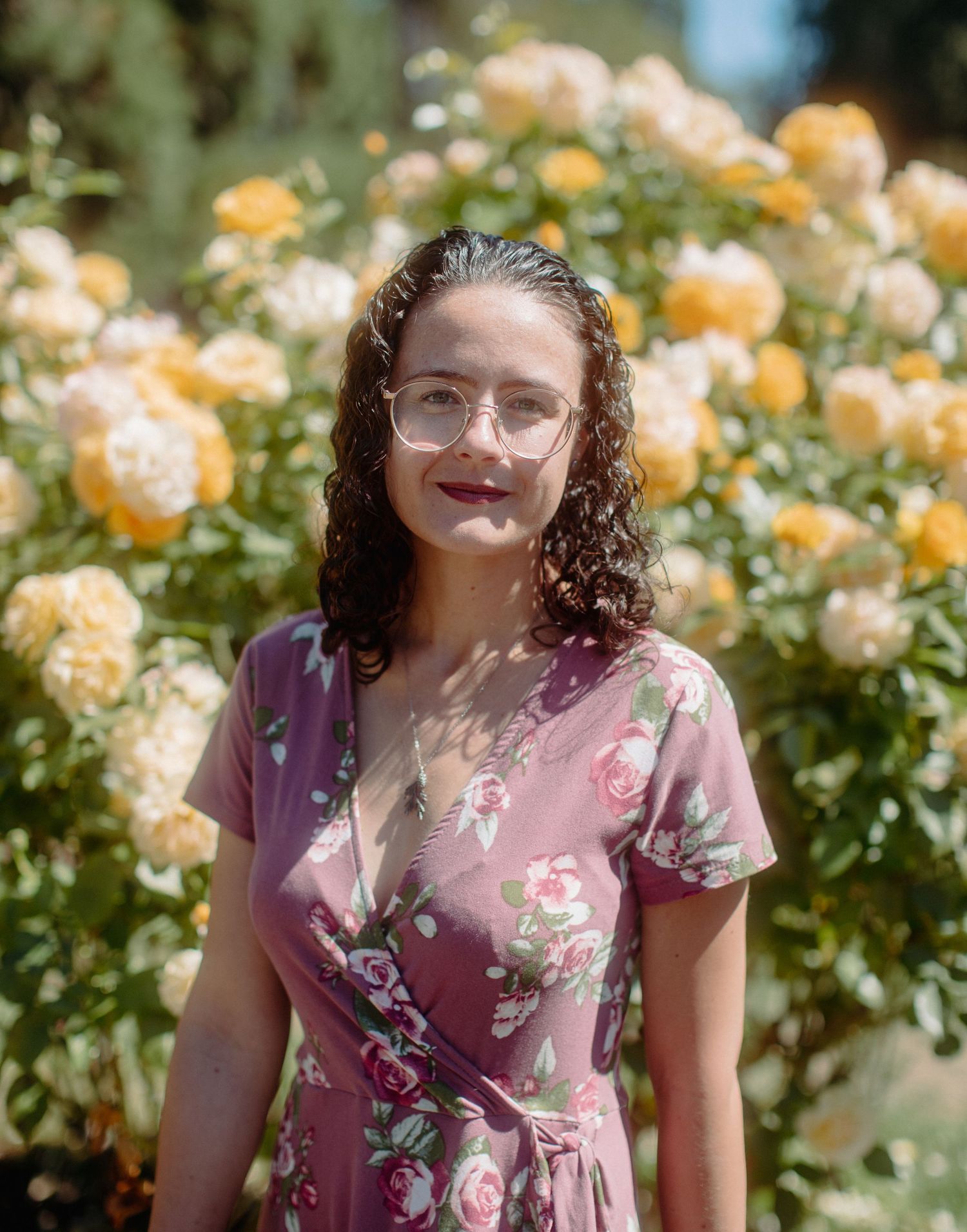 Young curly-haired woman in pink floral dress and glasses stands before yellow rose bushes.