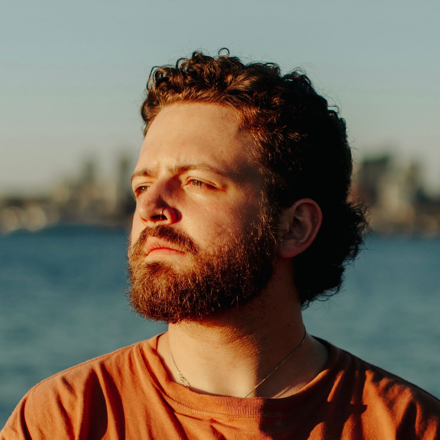 Bearded man with curly hair wearing an orange t-shirt stands near a waterfront in warm golden hour sunlight.