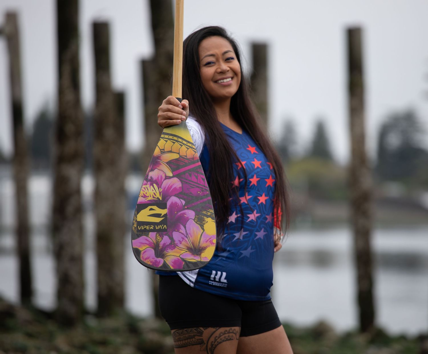 Smiling woman holds colorful paddle near waterfront with wooden posts in background.