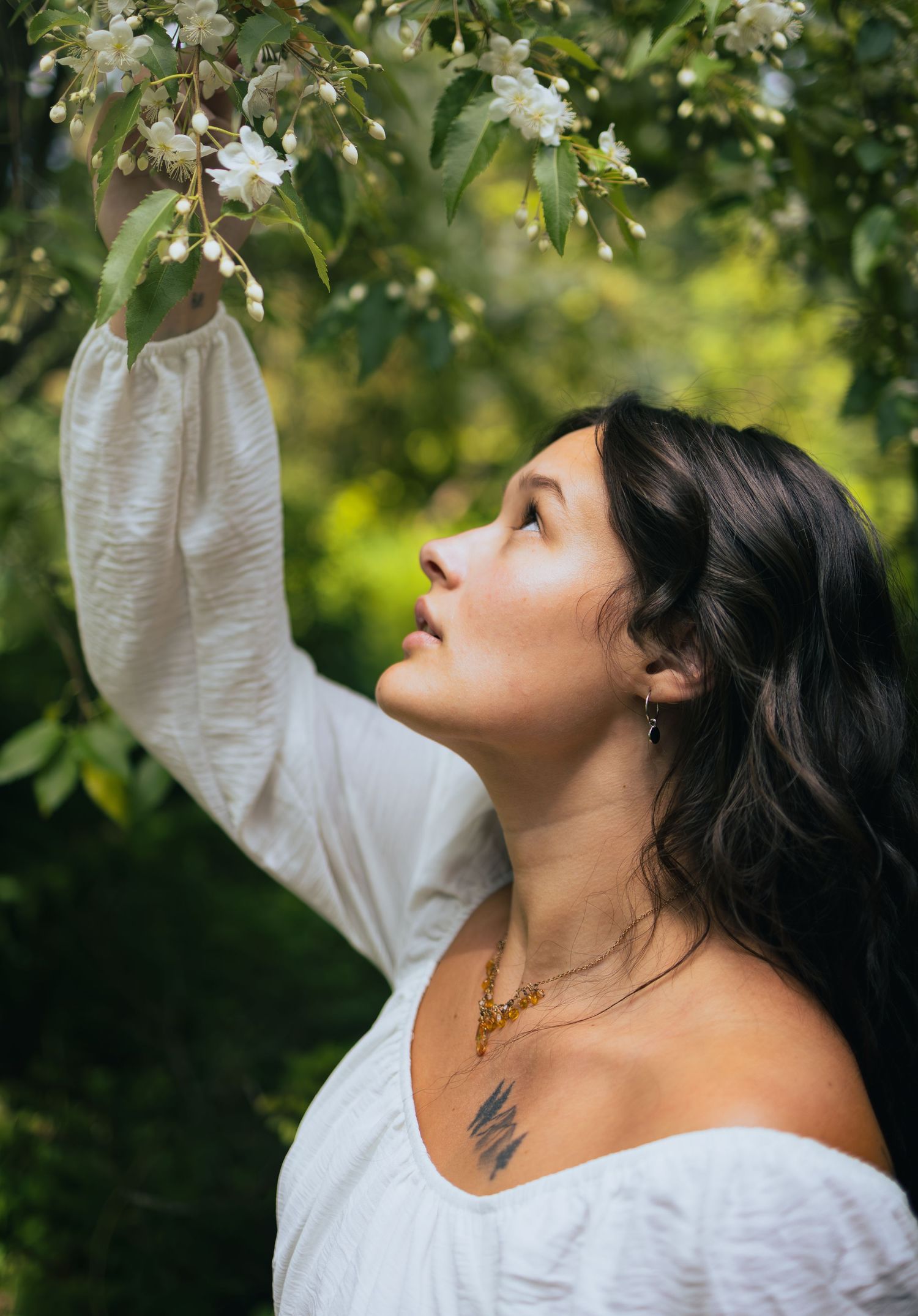 Woman in white off-shoulder top reaching up to touch white blossoms on a flowering tree in a lush green garden.