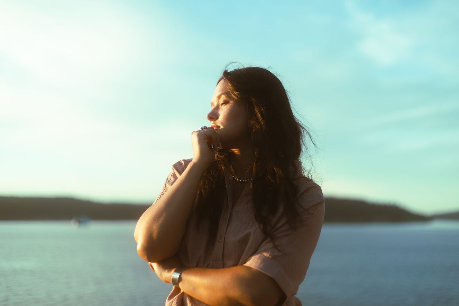 Young woman with long dark hair stands by water, eyes closed, basking in warm sunlight.