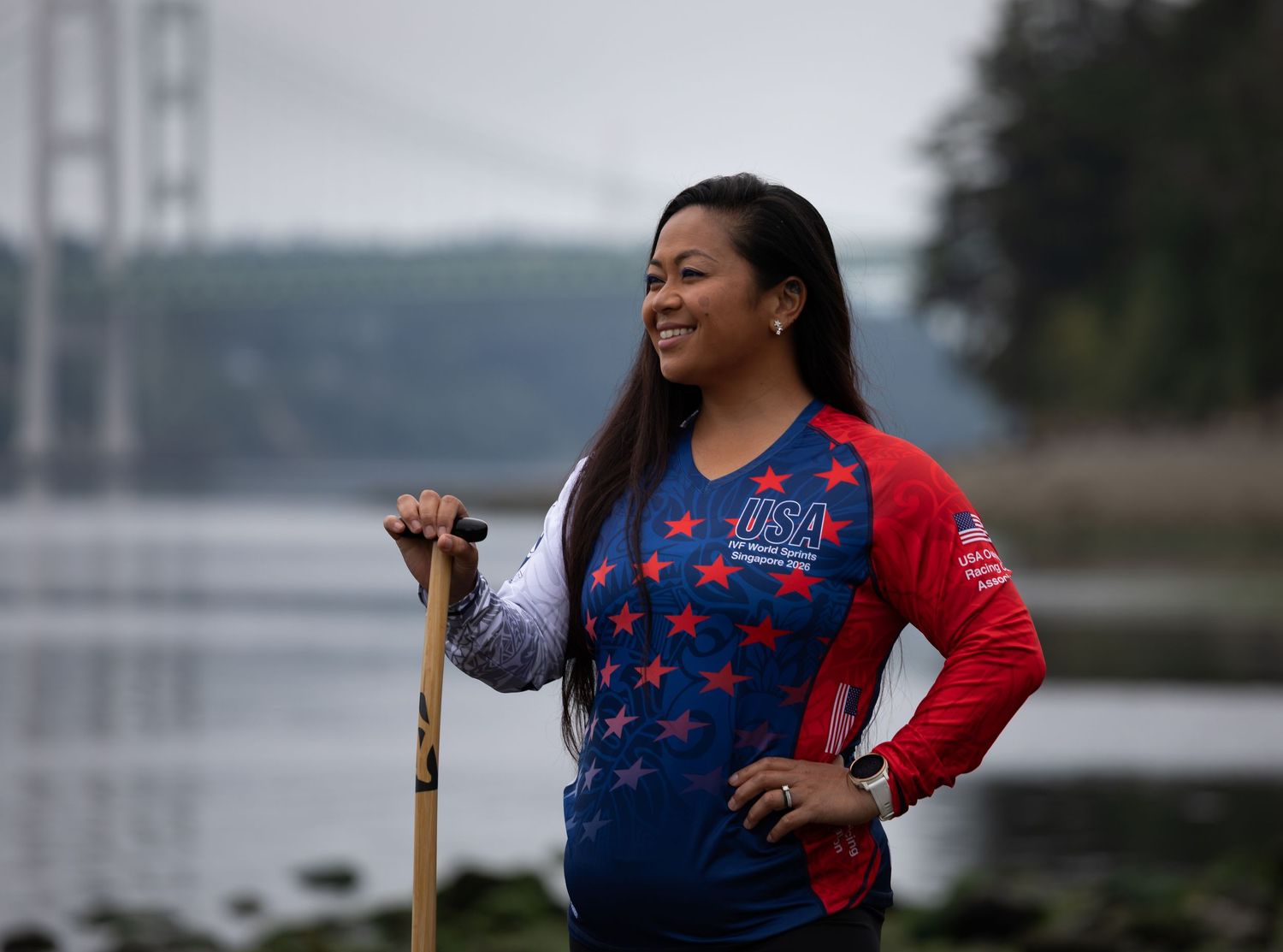 Female athlete in USA jersey stands confidently near a bridge and waterway holding a wooden paddle outdoors.