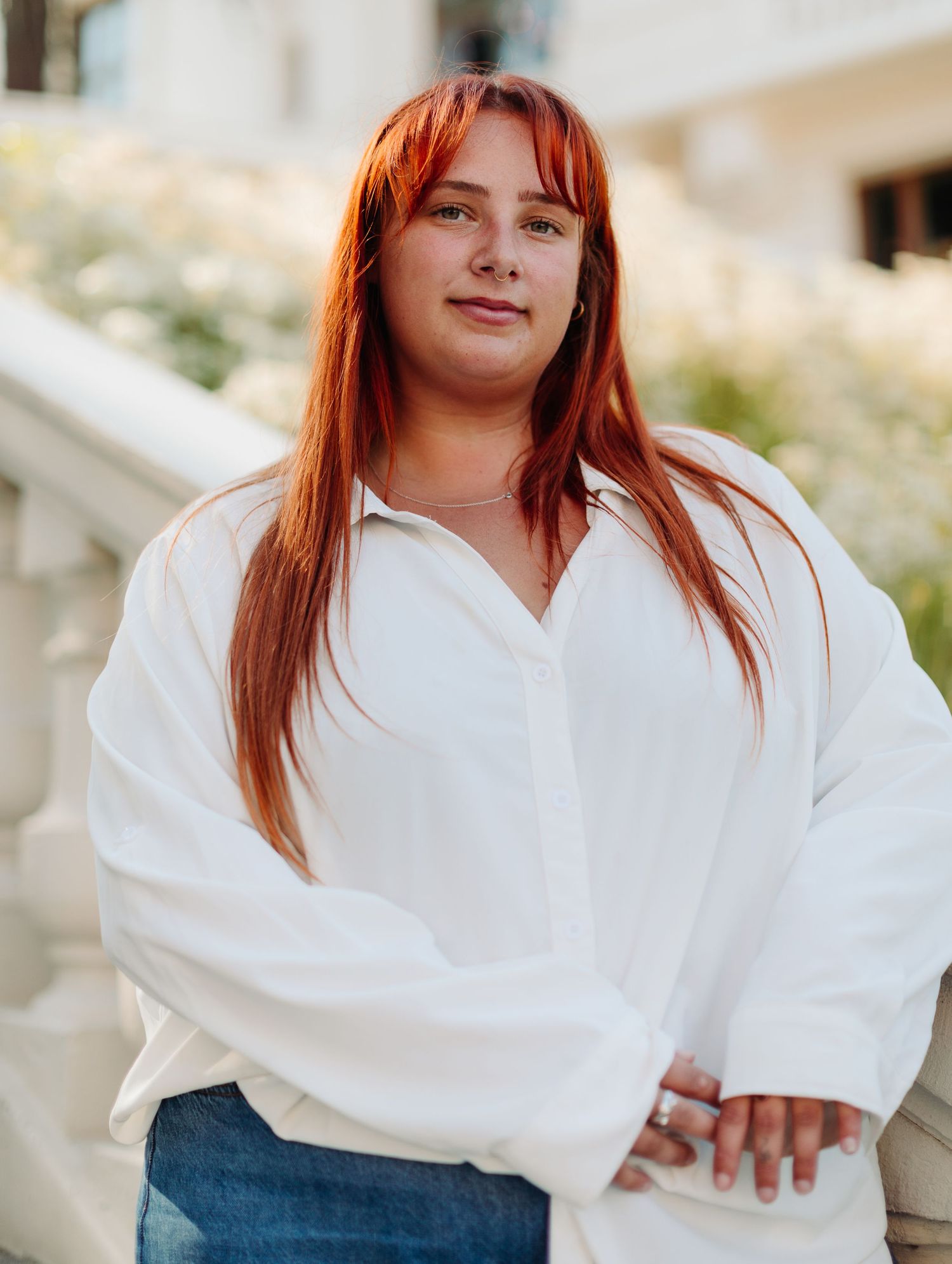 Young woman with red hair and bangs wearing white oversized shirt standing near white flowers.