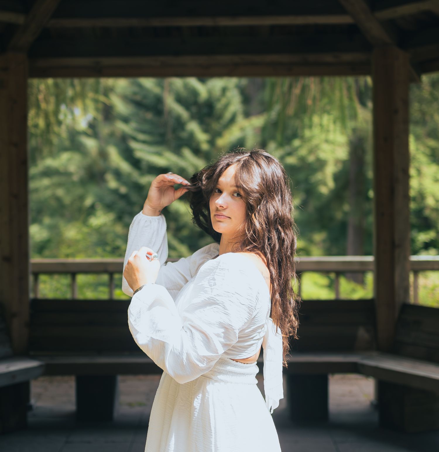 Woman in white long-sleeve dress posing inside wooden gazebo with evergreen trees in background.
