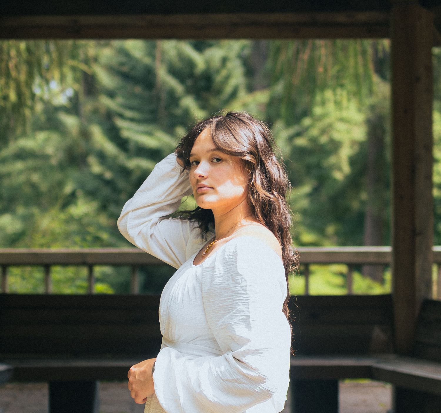 Woman in white long-sleeve dress posing under wooden pergola with pine trees in background.