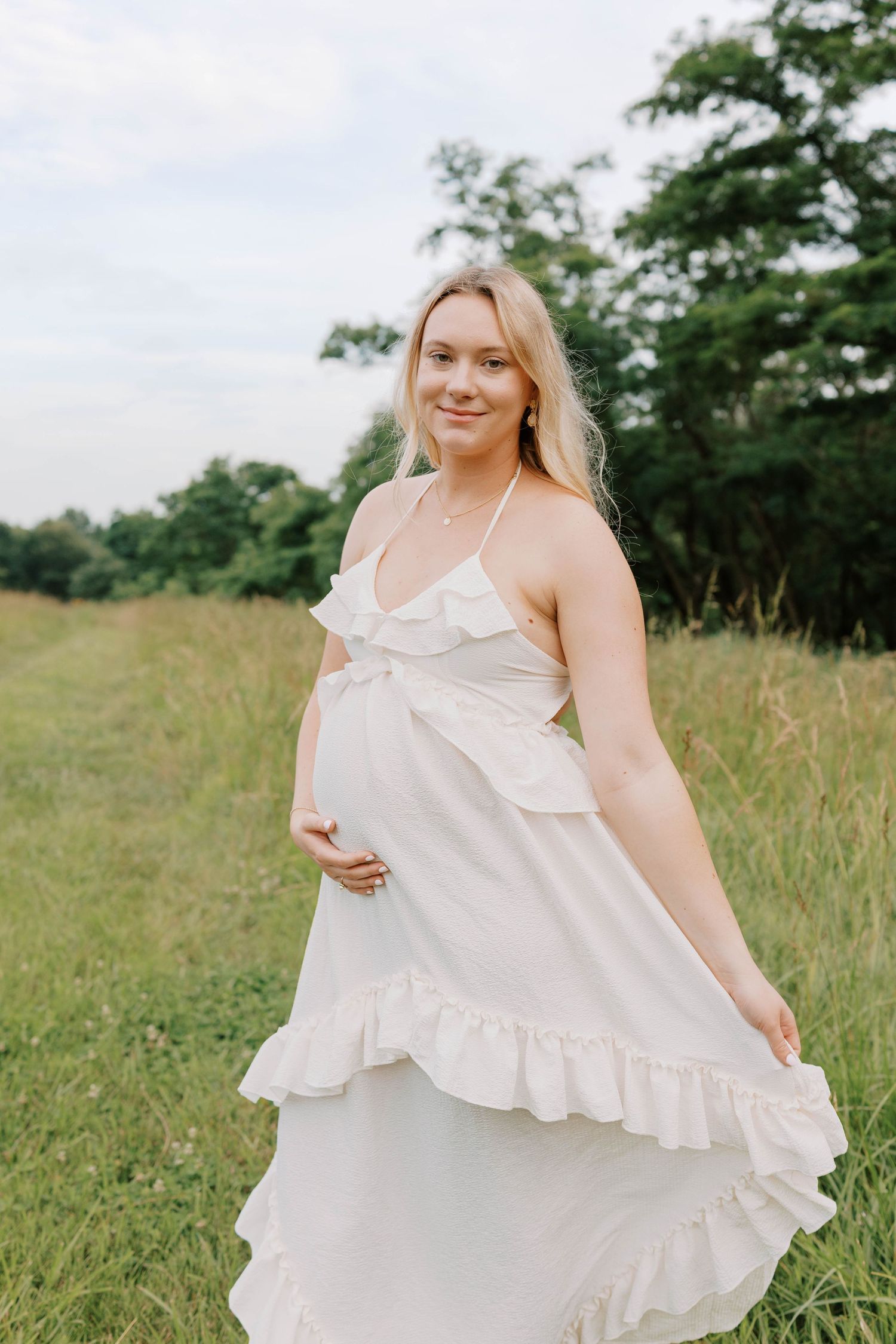 Smiling pregnant woman in white ruffled dress cradling baby bump in green field.