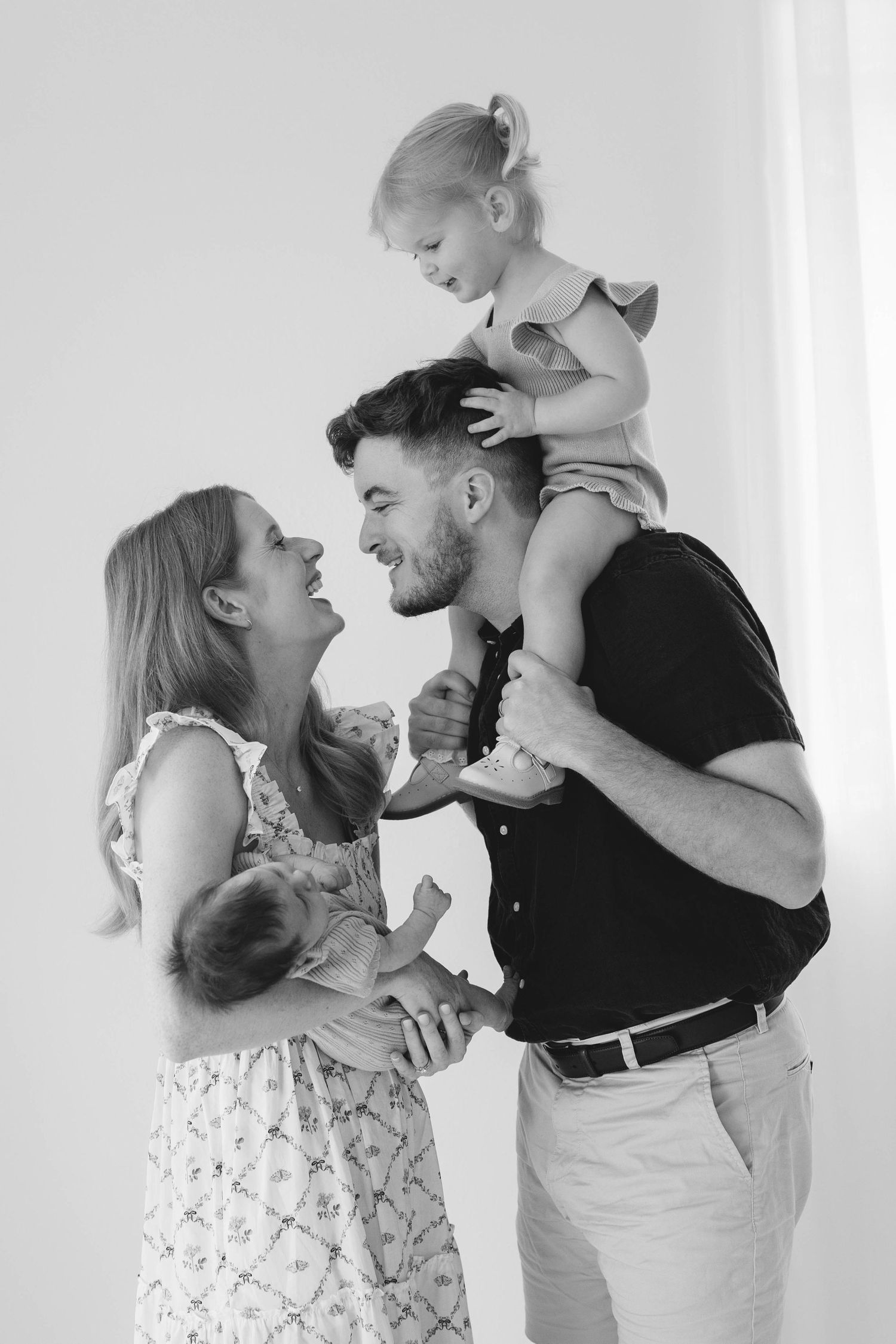 A black and white photo of a laughing family, with a toddler on dad's shoulders and mom holding a newborn.