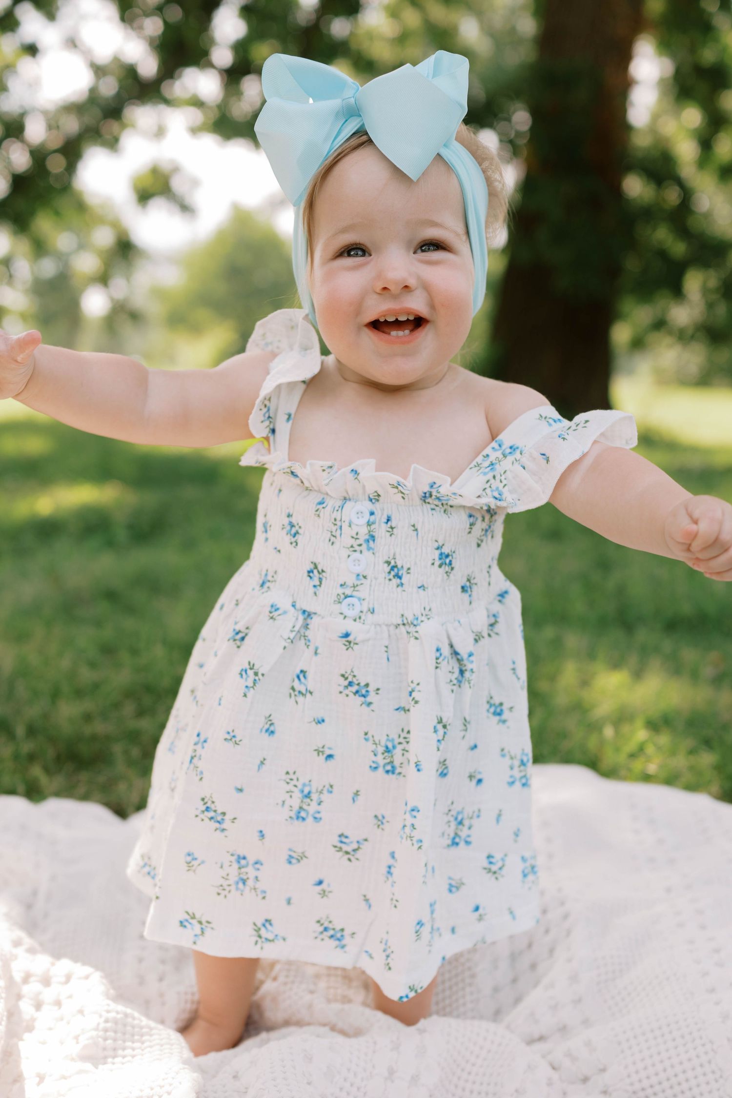 Happy toddler girl in a floral dress and blue bow headband standing on a white blanket outdoors.