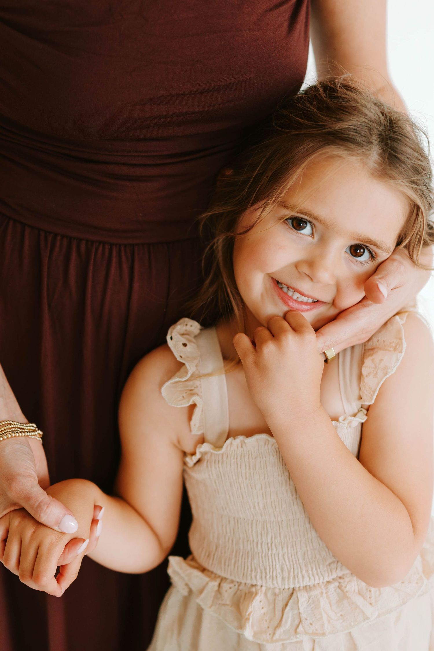 Adorable smiling little girl in a cream dress leans against her mother's leg holding her hand in all white studio in Kirkwood, Missouri.