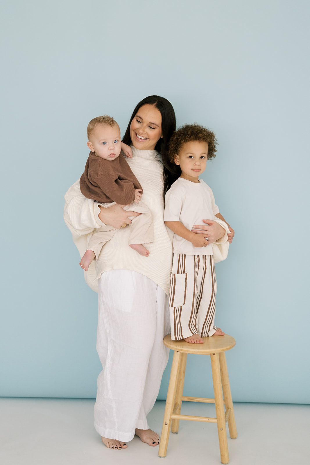 A joyful mother in white holds her infant while her curly-haired toddler stands on a stool beside her.