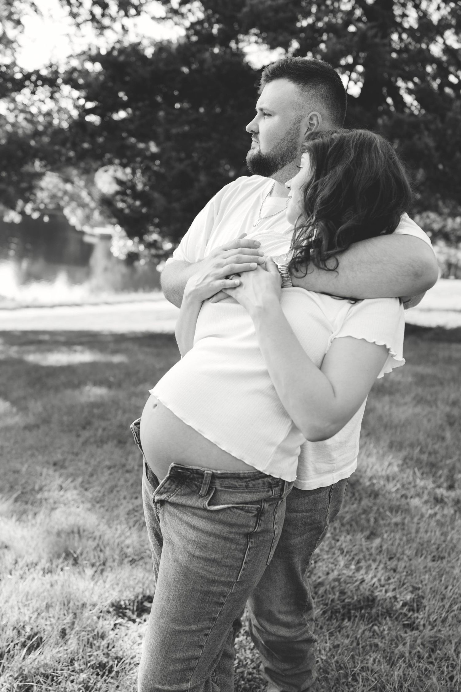 Black and white photo of pregnant couple embracing tenderly outdoors under trees.