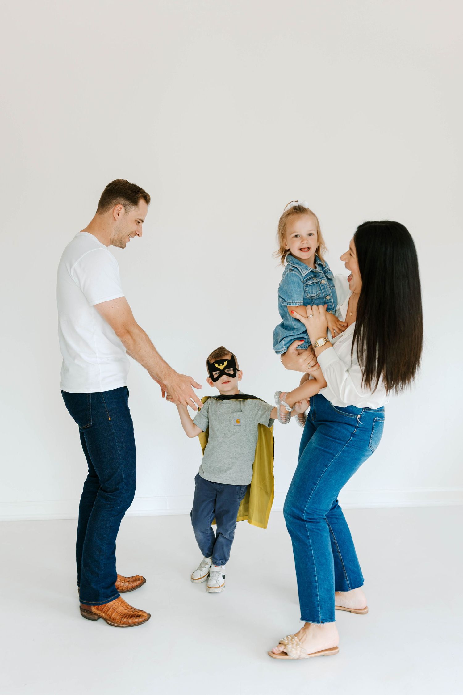 A joyful family plays together as a boy in a superhero costume stands between his parents holding a baby girl.