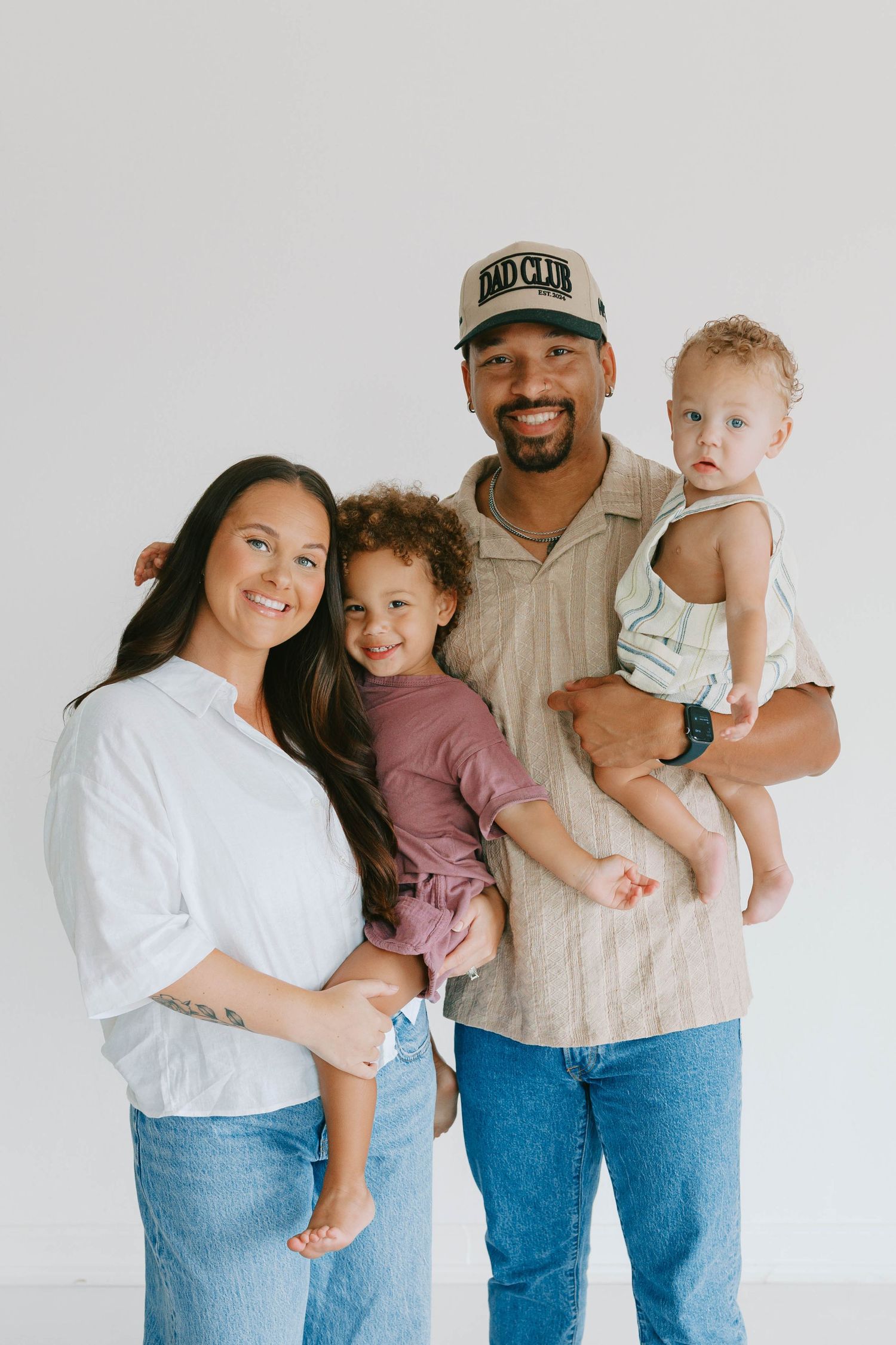 Smiling mixed-race family of four poses in a bright white studio, parents each holding a young child.