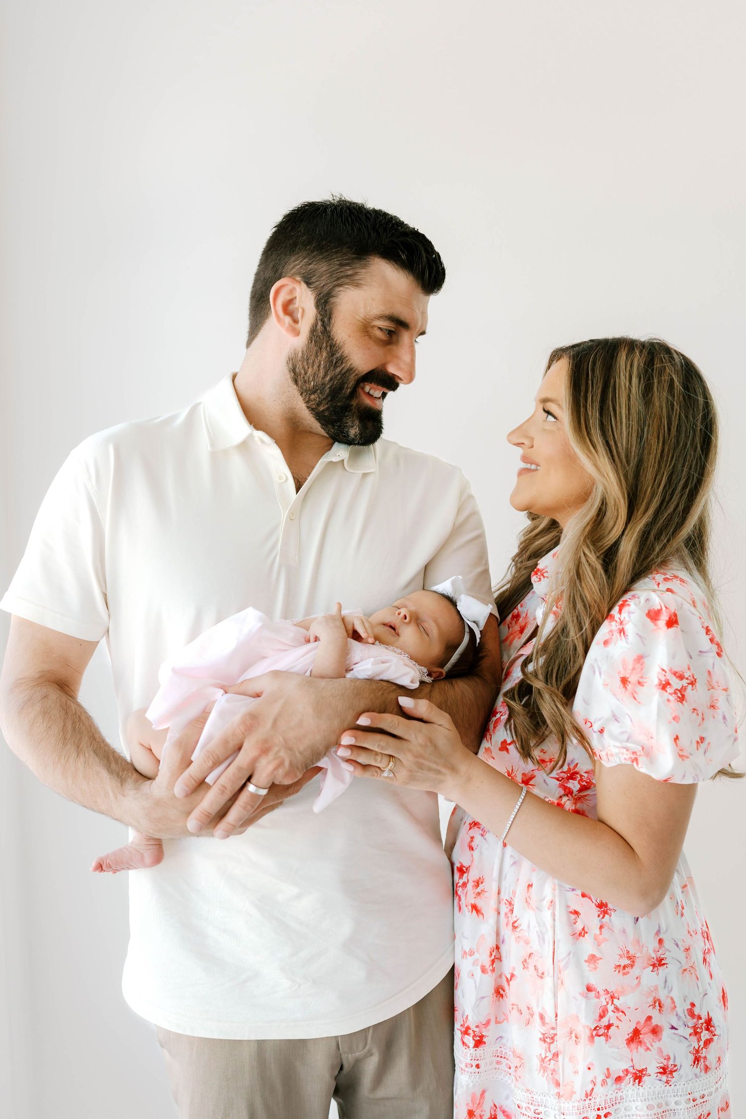 Smiling parents in white outfits lovingly hold their newborn baby girl in a bright studio setting.