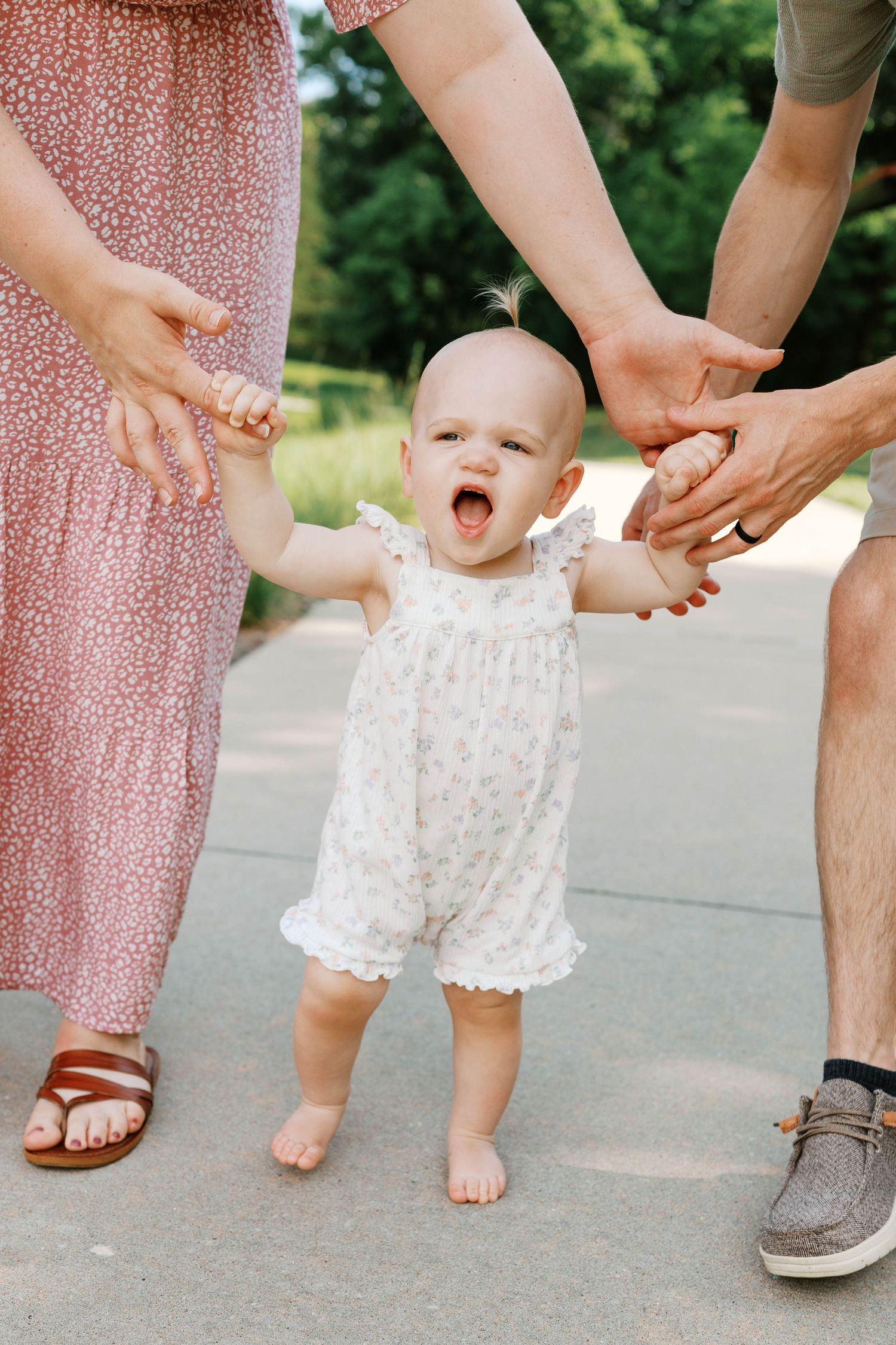 Baby in floral romper taking first steps while parents hold hands outdoors in park.