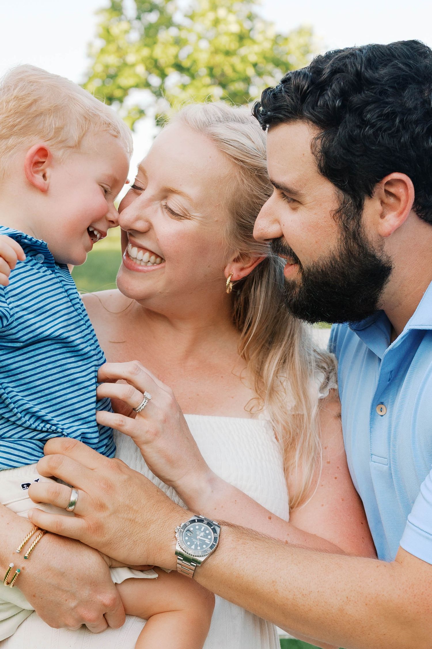 Happy young family with smiling toddler boy being held by laughing parents outdoors.