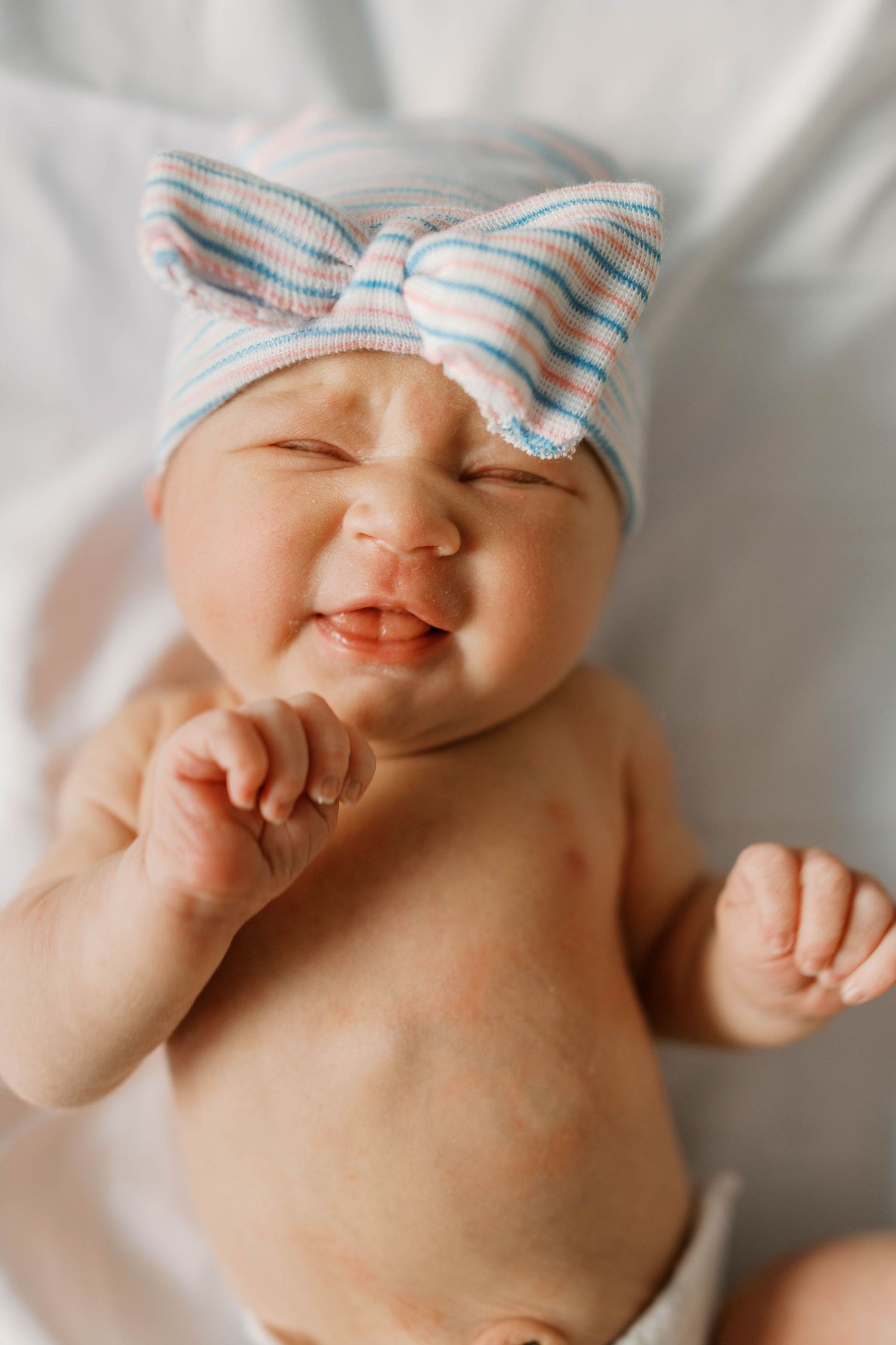 A happy newborn baby in a striped bow hospital hat smiling with eyes closed on white blanket.