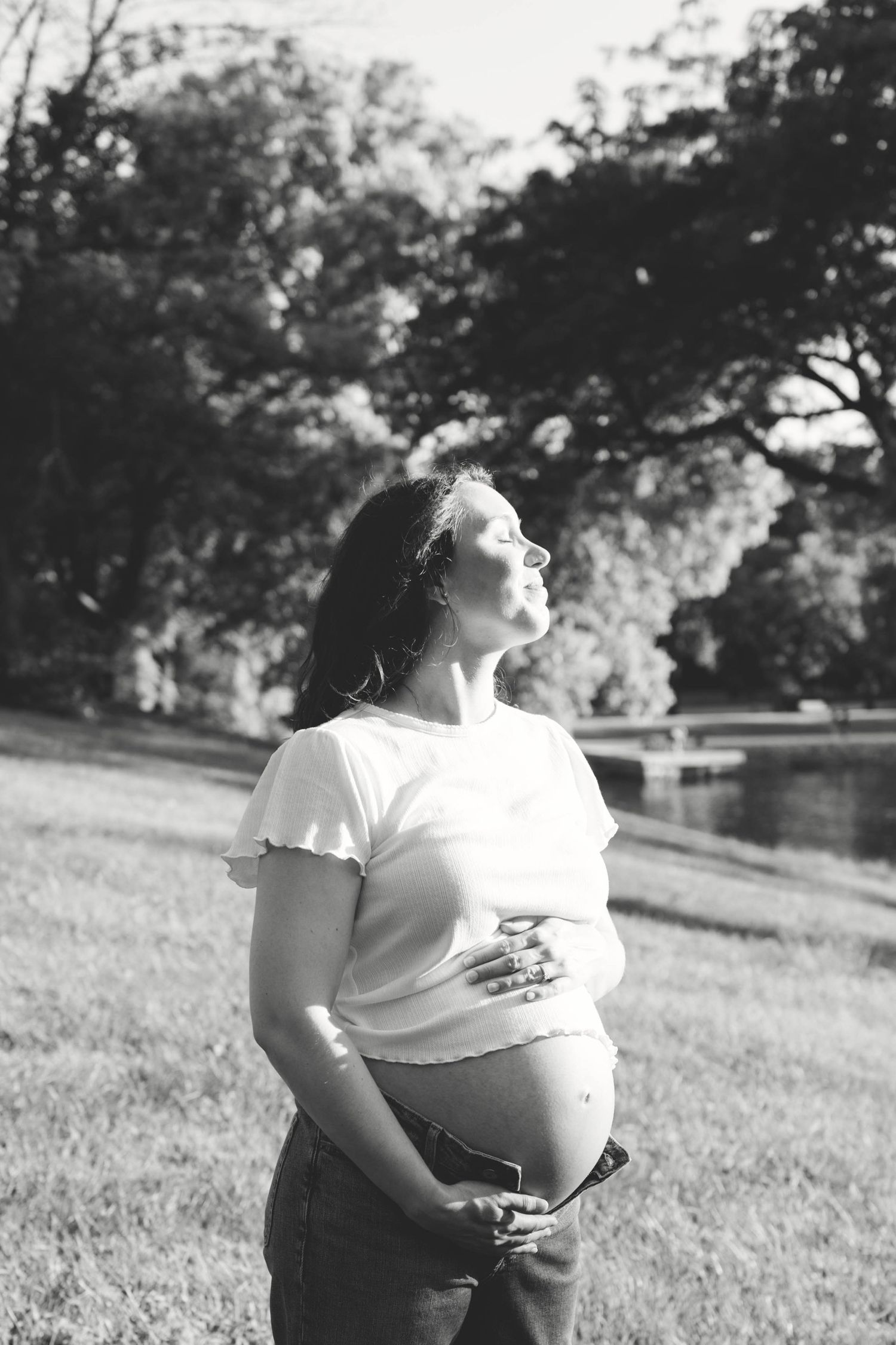 Black and white portrait of pregnant woman holding belly while standing in sunny park.